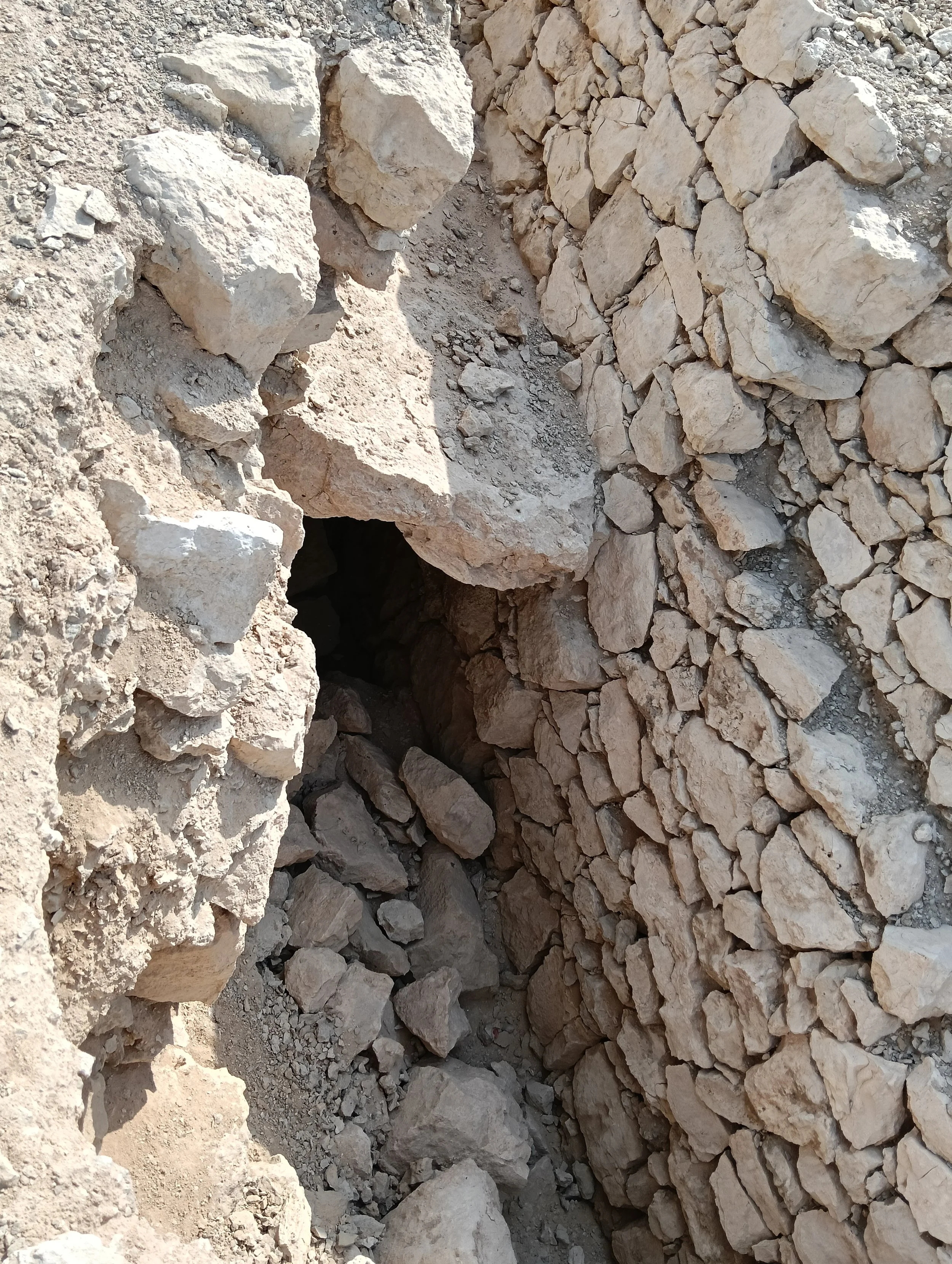 Looking down into a narrow stone-lined opening, with a large slab spanning a dark recess and rubble below.