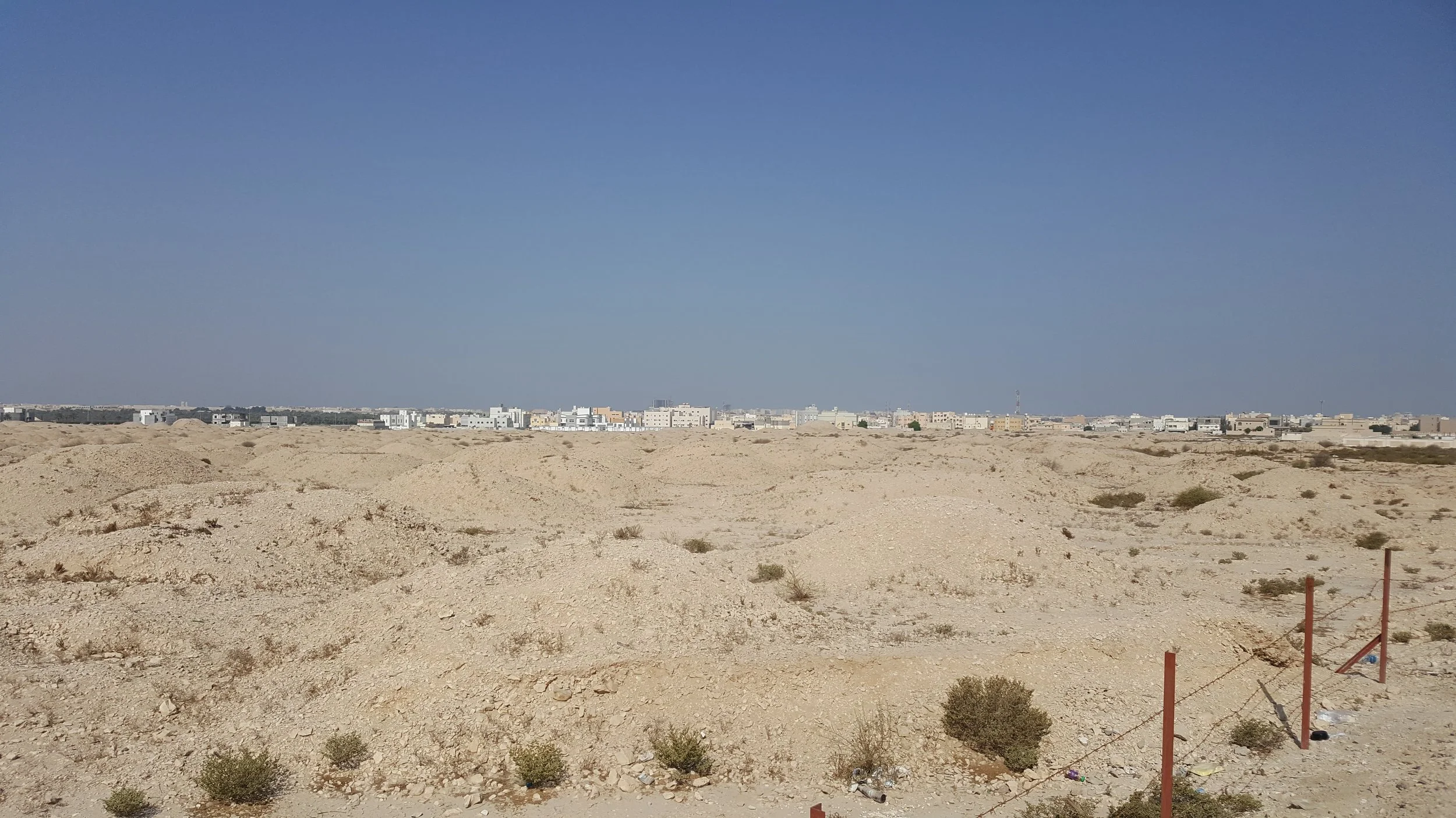 A wire fence and red-brown posts cross the edge of a broad mound landscape beneath modern houses on the horizon.