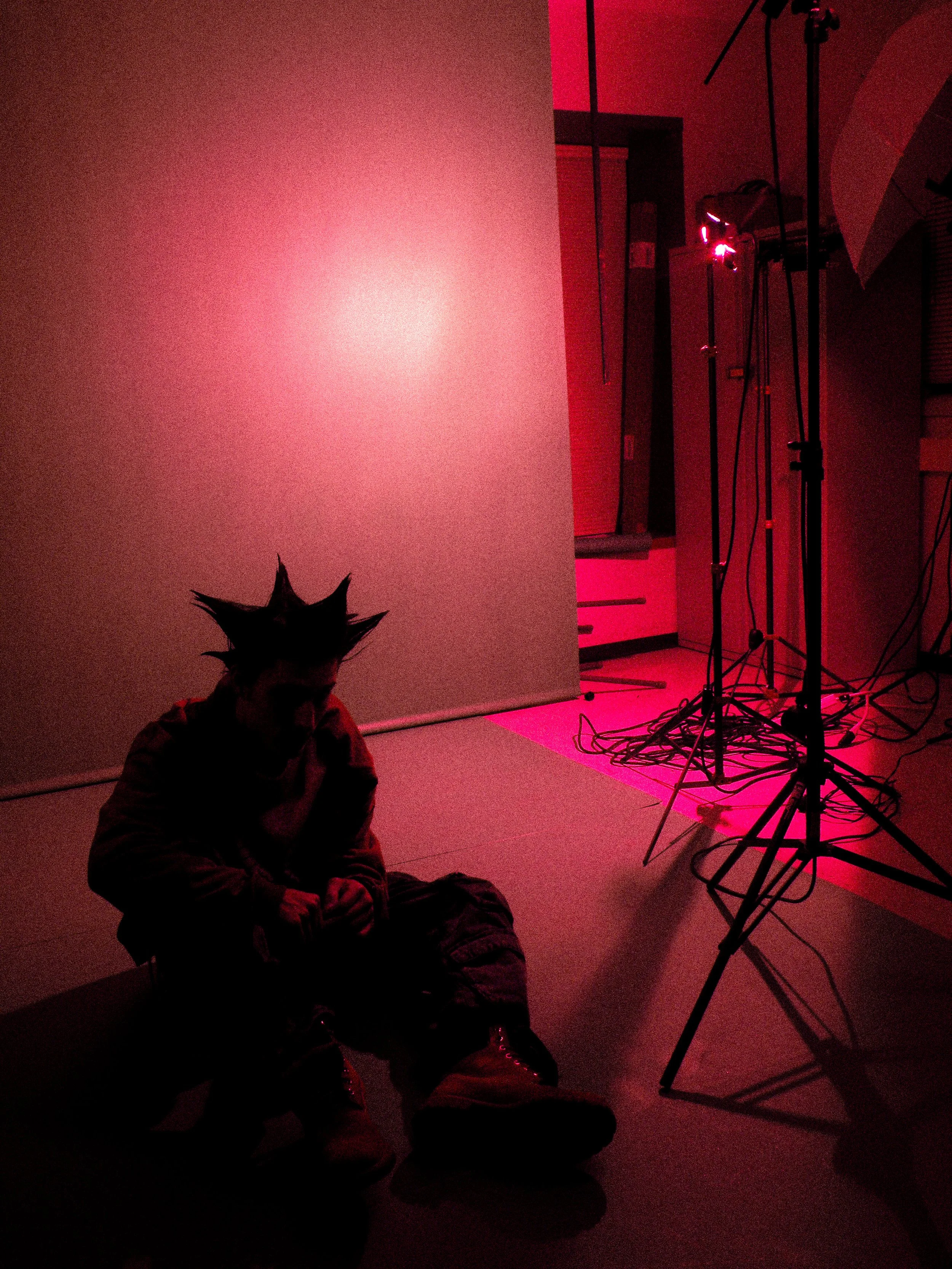 A young man with spiky hair sitting on the floor in front of a pink lighting setup, with studio lights and tangled cables in a photography studio.