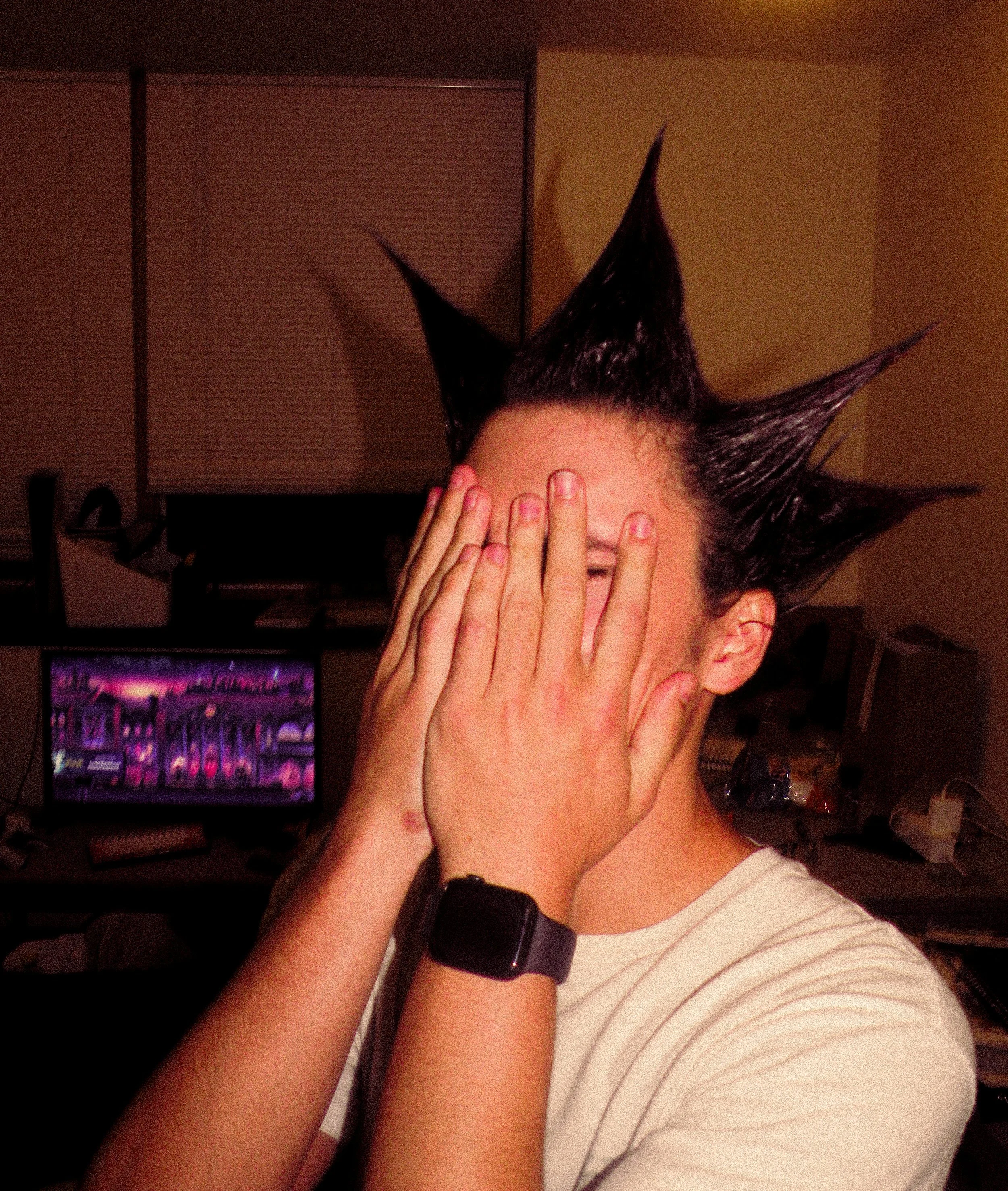 Person with spiked hair covering their face with both hands, wearing a wristband, in a room with a TV and various objects.