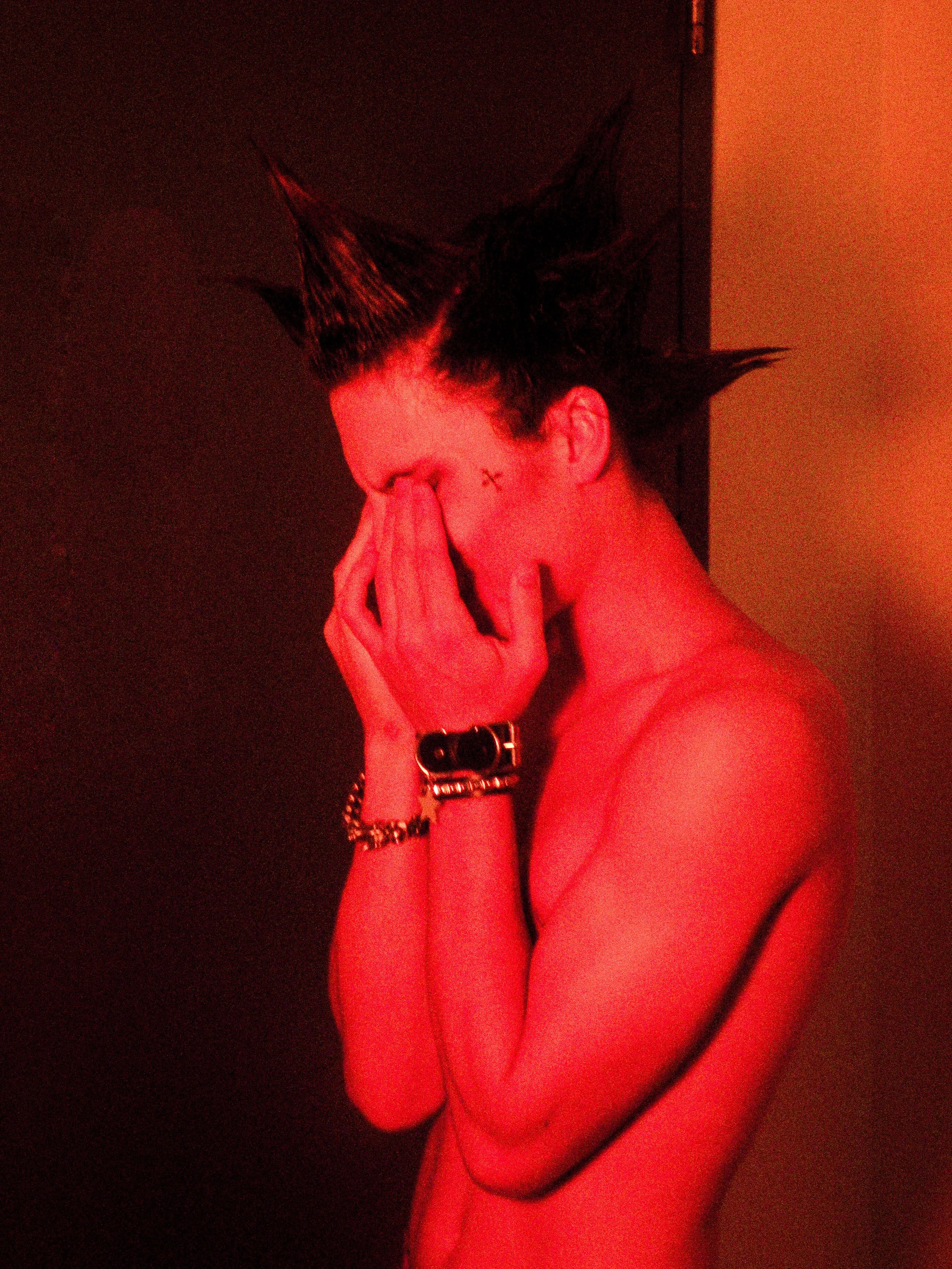 Person with spiky hair covering face with hands, painted in red tint.