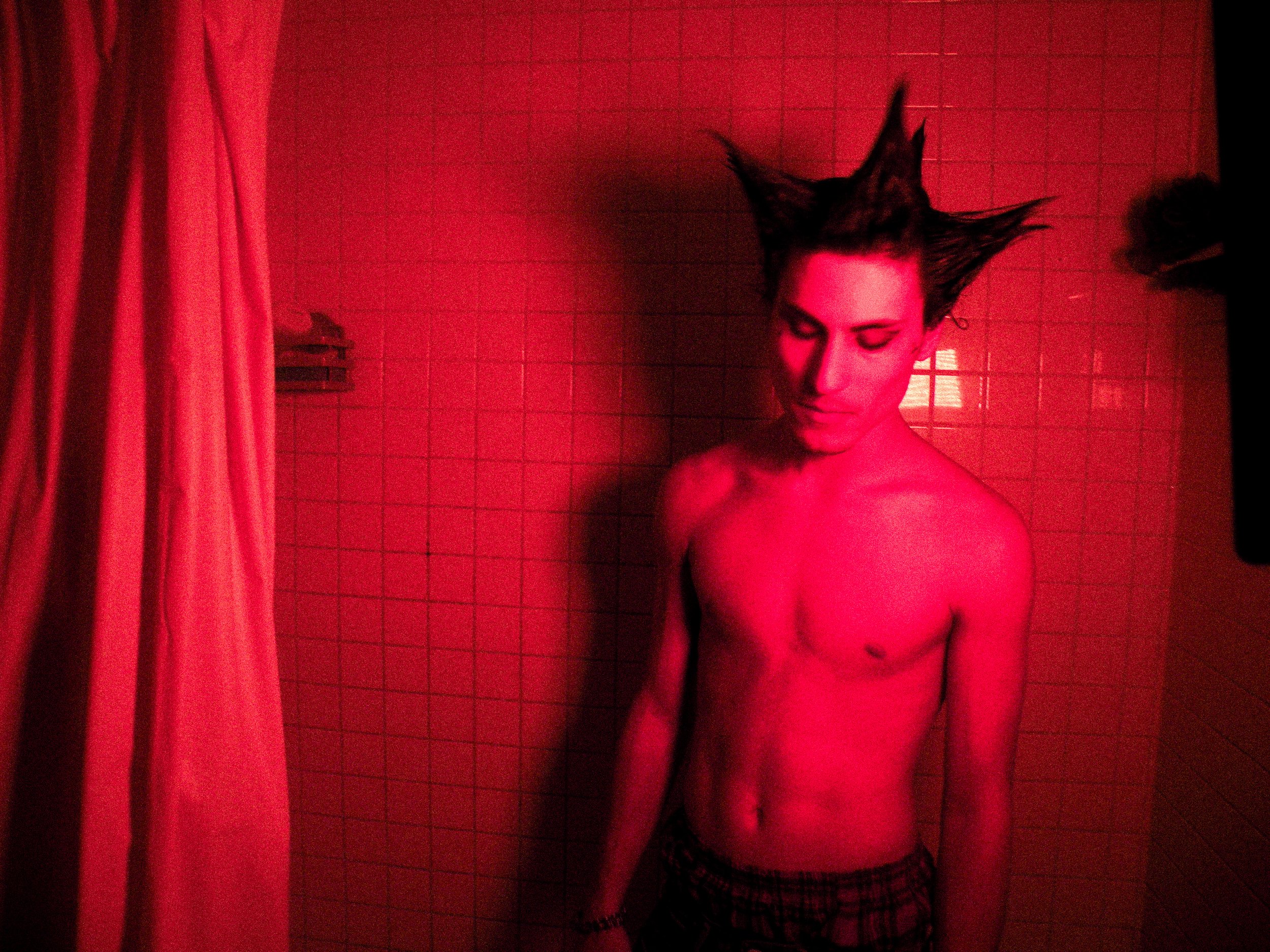 A young man with spiky hair standing in a shower, illuminated by red light, with his eyes closed and head slightly bowed.
