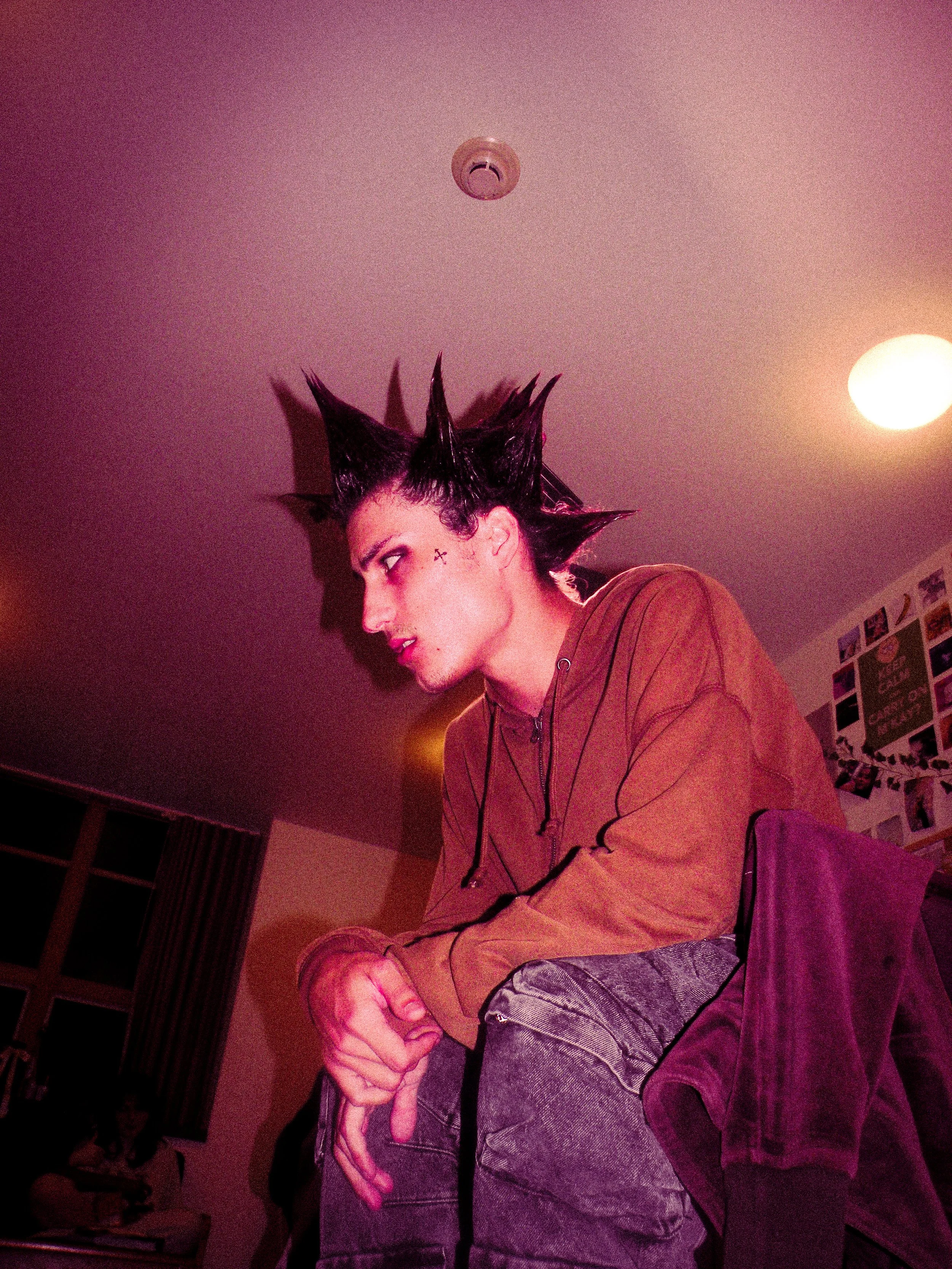 A person with spiky, styled hair and a small cross tattoo near the eye, wearing a brown hoodie and jeans, sitting indoors with a serious expression, under pinkish and yellow lighting.