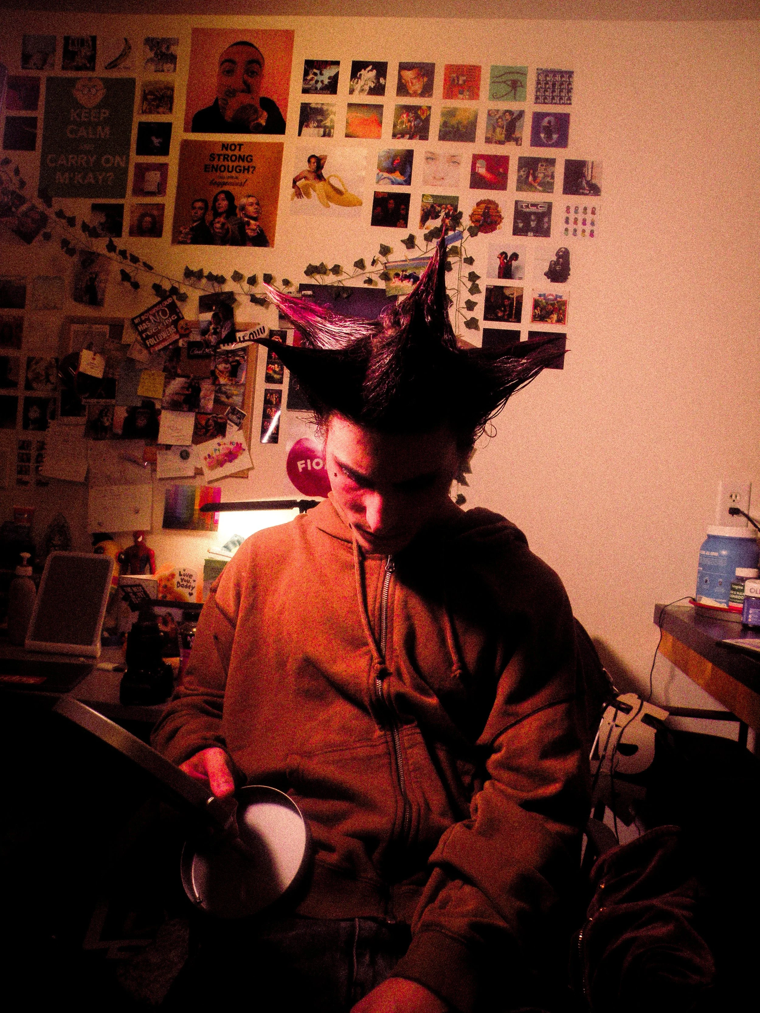 A person with spiky hair is sitting in a dimly lit room, looking down at a tablet device. The room has various posters, photos, and decorations on the wall behind them, including motivational quotes and pictures of people and scenes.