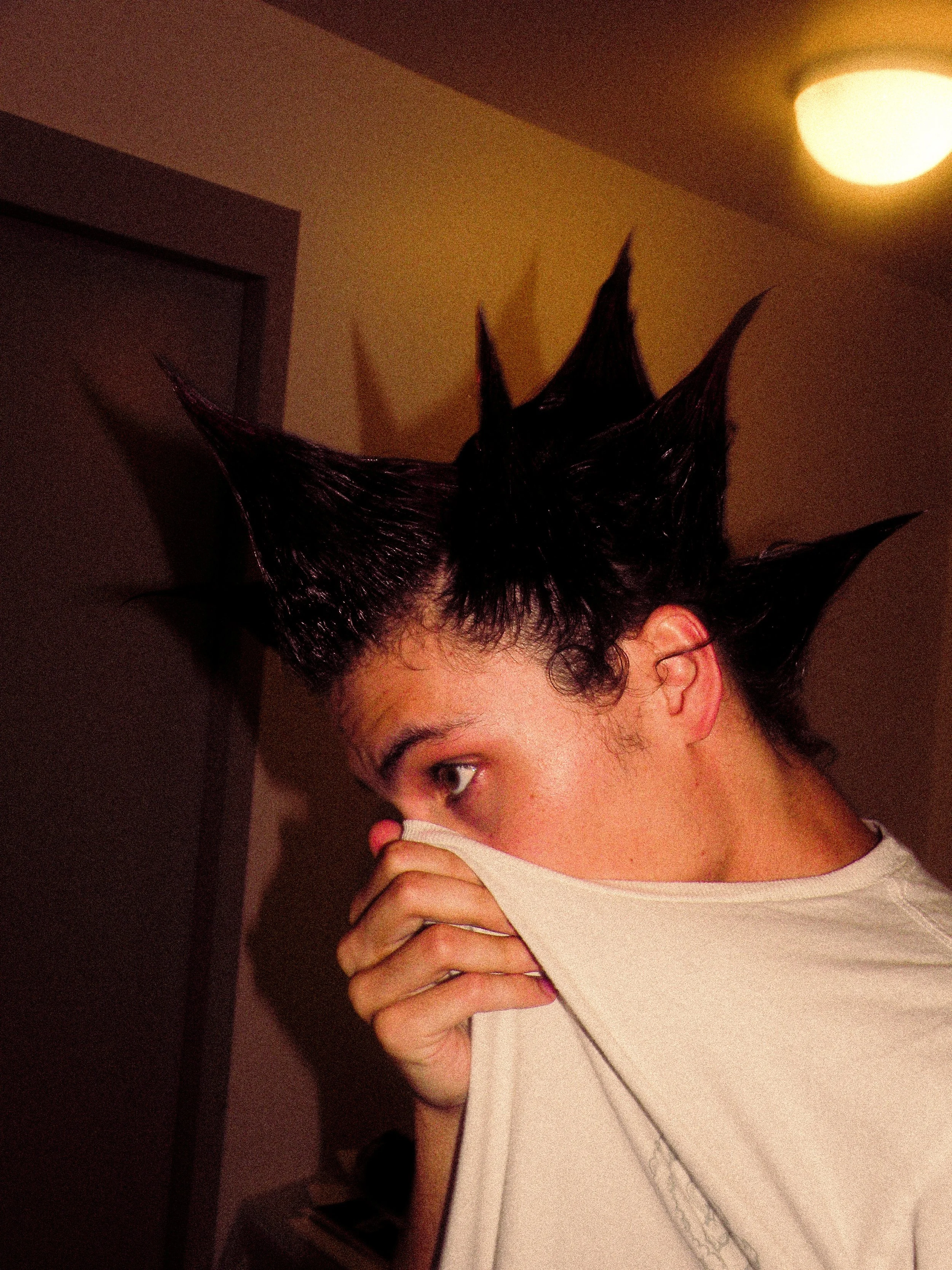 Person with spiked, styled black hair covering their mouth with a white T-shirt, in an indoor setting.
