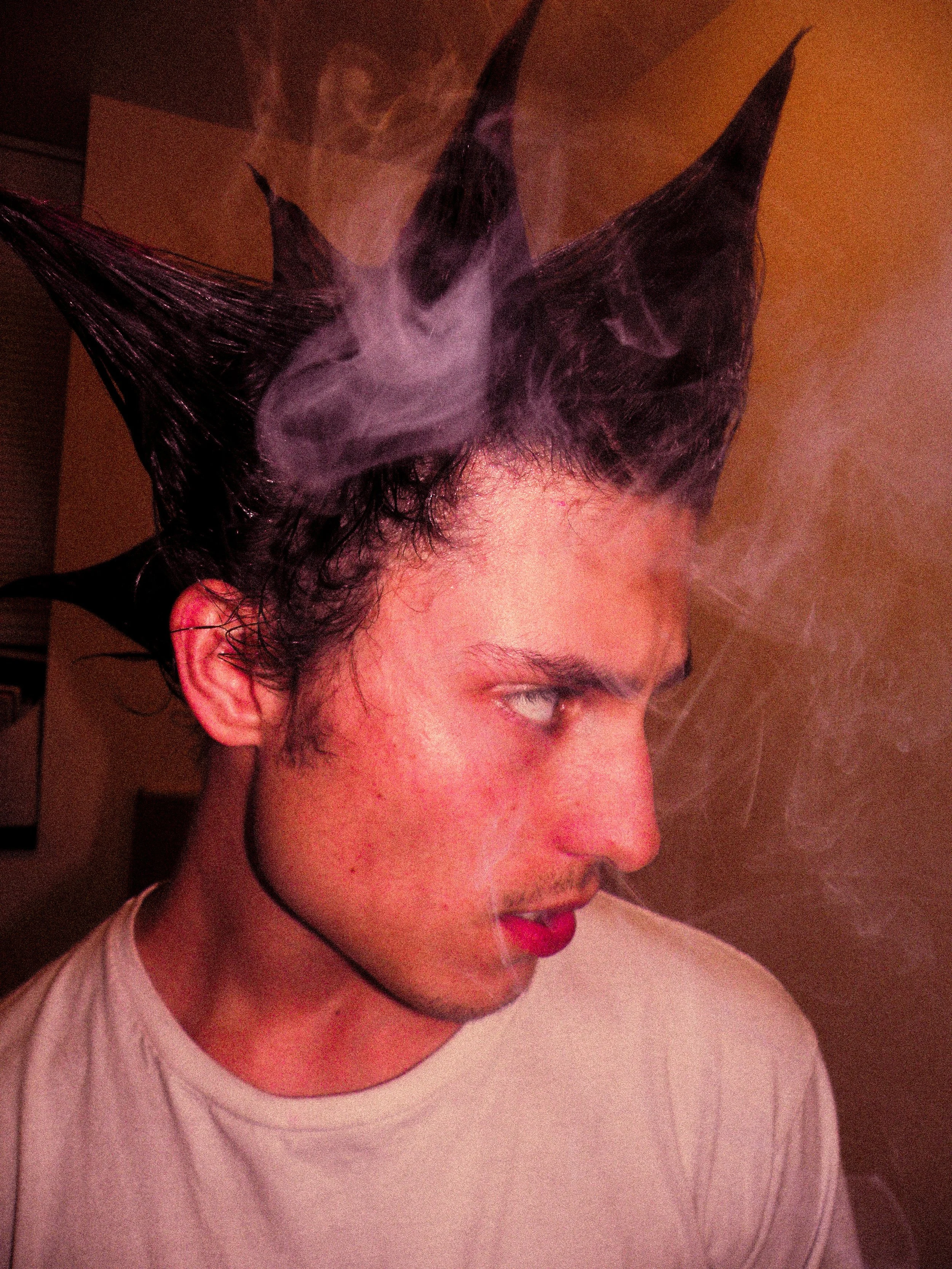A young man with dark, spiked hair, and makeup, exhaling smoke.