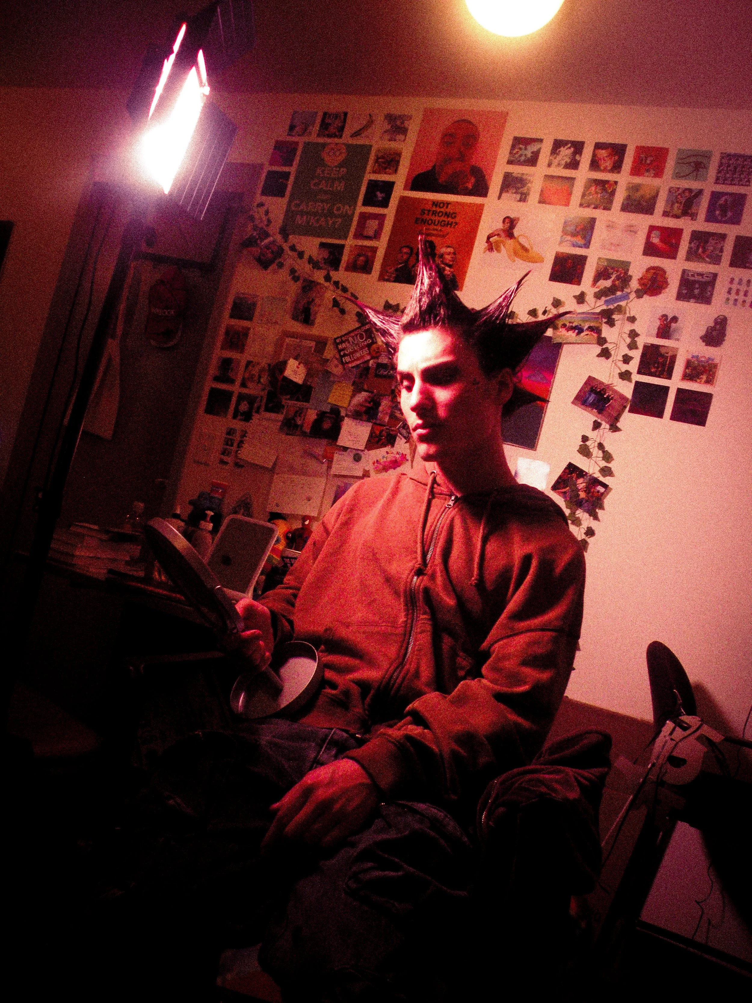 A person with spiked hair seated in a dimly lit room with posters and photos on the wall behind them, holding a tablet.