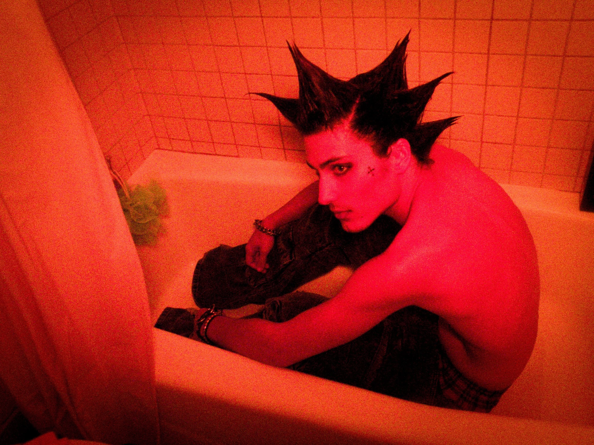 Person with spiked hair and makeup sitting in a bathtub with a red tint, looking over their shoulder.