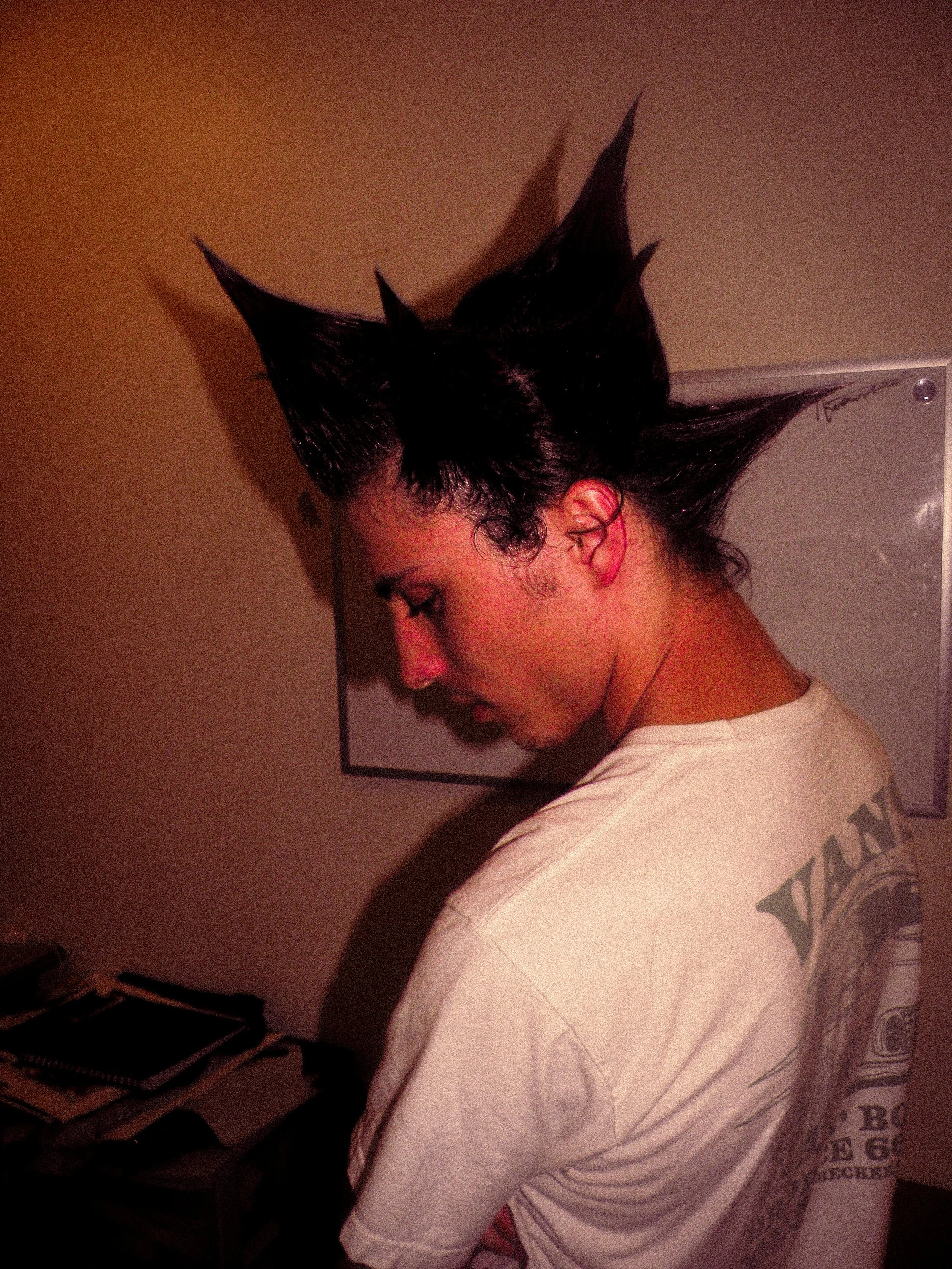 Person with styled spiky black hair, standing sideways, wearing a white t-shirt, indoors near a wall with a framed picture and a table with books.