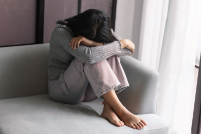 A woman sitting on a gray sofa near a window, with her head resting on her knees and her arms covering her face, appearing upset or sad.