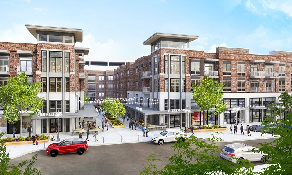 A modern mixed-use urban development with brick buildings, retail stores on the ground floor, and residential units above. Pedestrians walk along the wide sidewalk, and cars are parked and driving on the street.