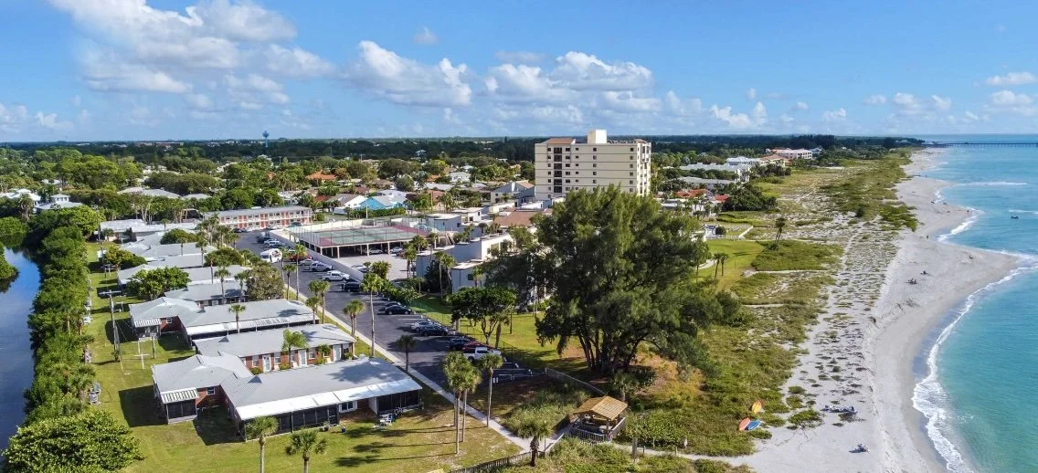 Aerial view of a coastal town with houses, trees, a river, a beach, and the ocean under a partly cloudy sky.