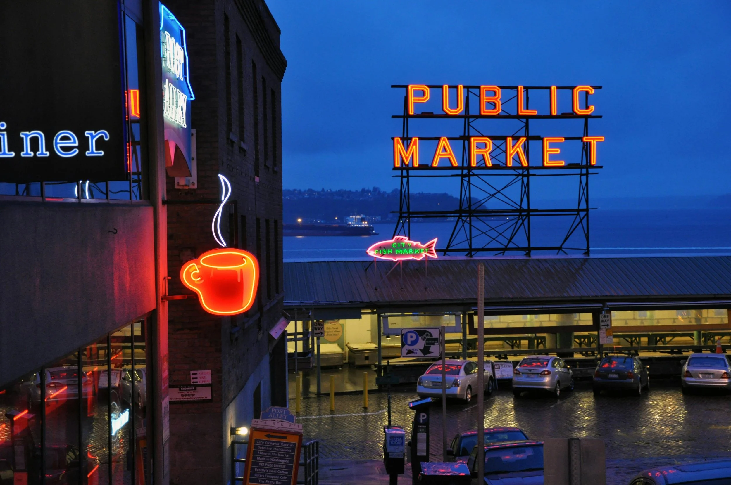 Neon signs outside a public market in Seattle, including a coffee mug, a fish, and the words 'Public Market', with a parking lot and a body of water in the background at dusk.