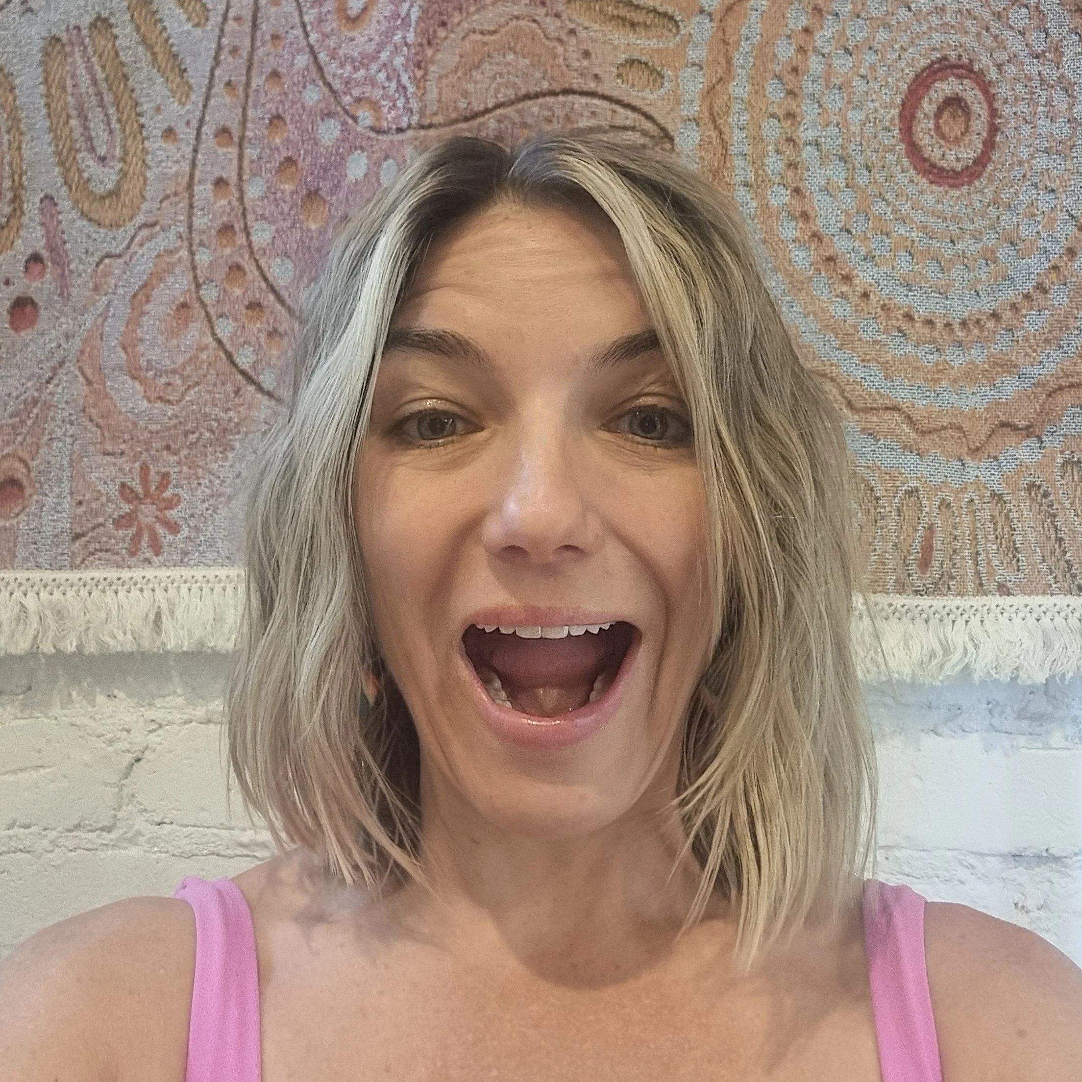 A woman with shoulder-length blonde hair smiling and opening her mouth, wearing a pink tank top, with a colorful tapestry hanging on a white brick wall behind her.