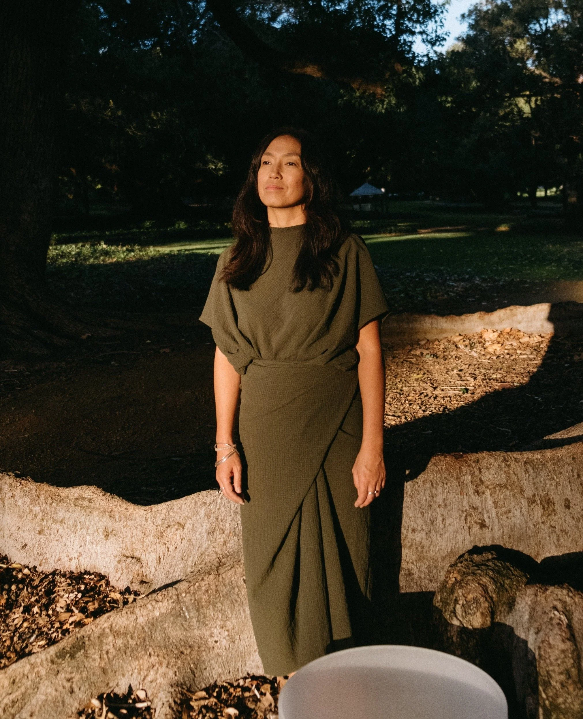 A woman in an olive green dress stands outdoors at night near a large tree, illuminated by an external light, with greenery and a small pavilion visible in the background.