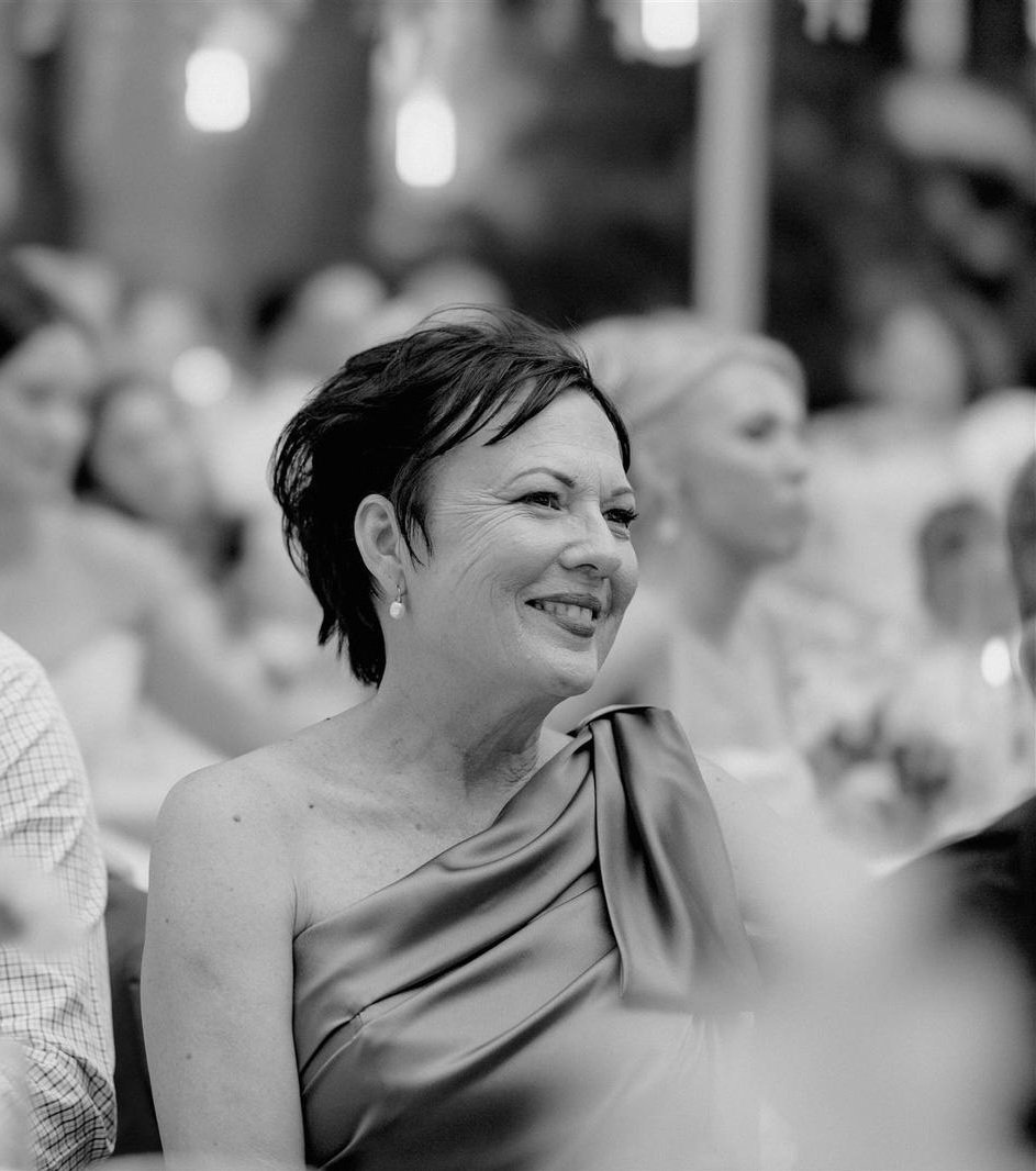 A woman with short dark hair and earrings wearing a strapless satin dress, smiling at an indoor formal event, with other women in the background.