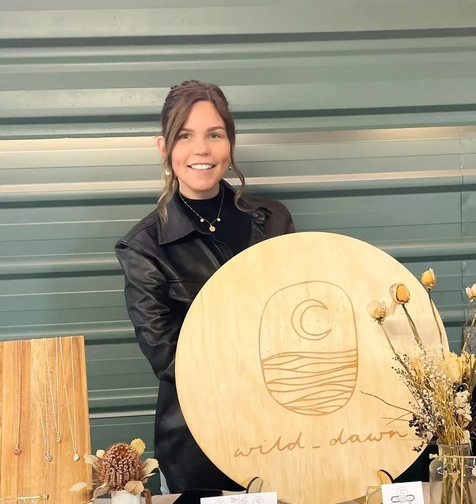 A woman with brown hair wearing a black leather jacket, smiling and standing behind a display table with jewelry and dried flowers, holding a large wooden sign that has a moon and waves design with the words 'wild dawn' written underneath.