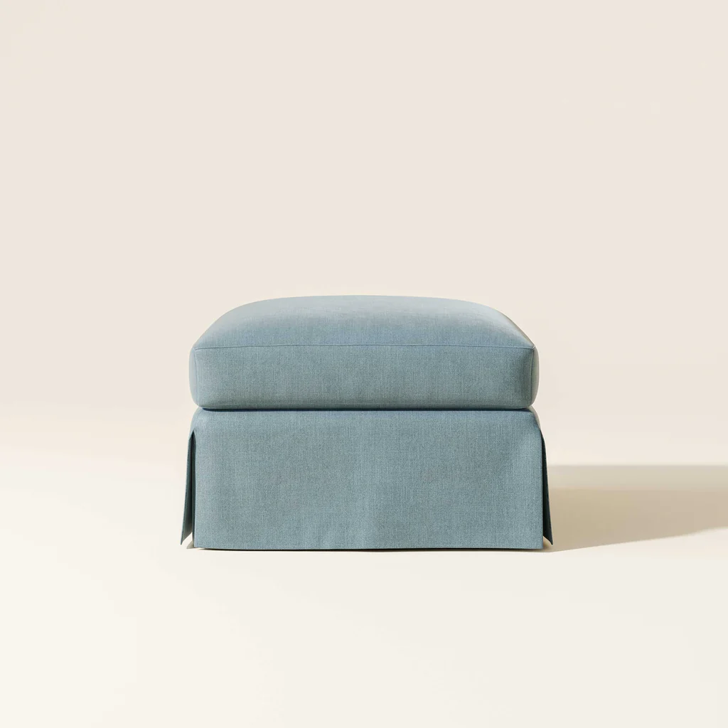 Light blue upholstered ottoman with fabric skirt on beige background.