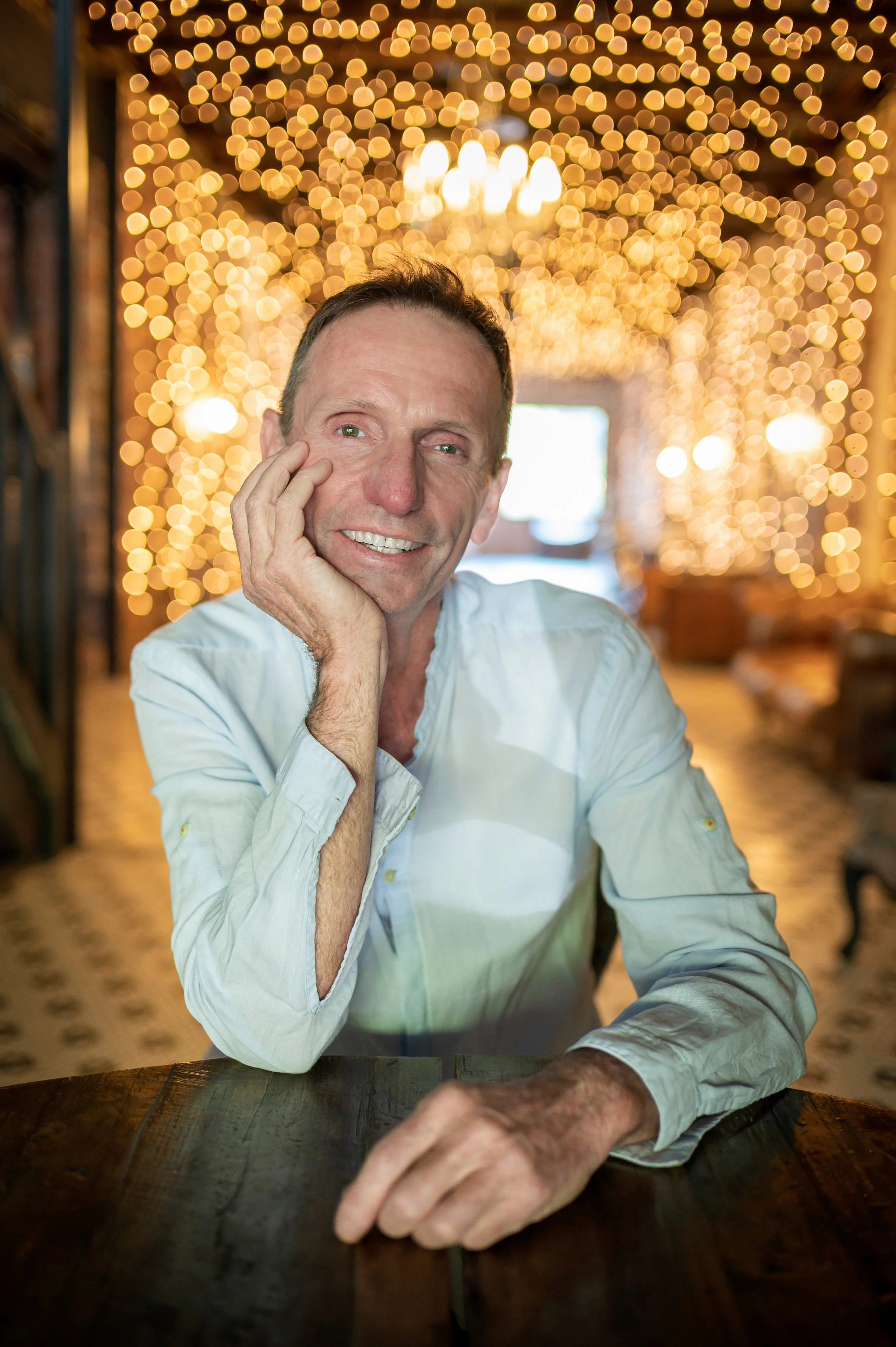 Jaye Stevens in a white shirt sitting at a wooden table with a warm, bokeh-lit background.