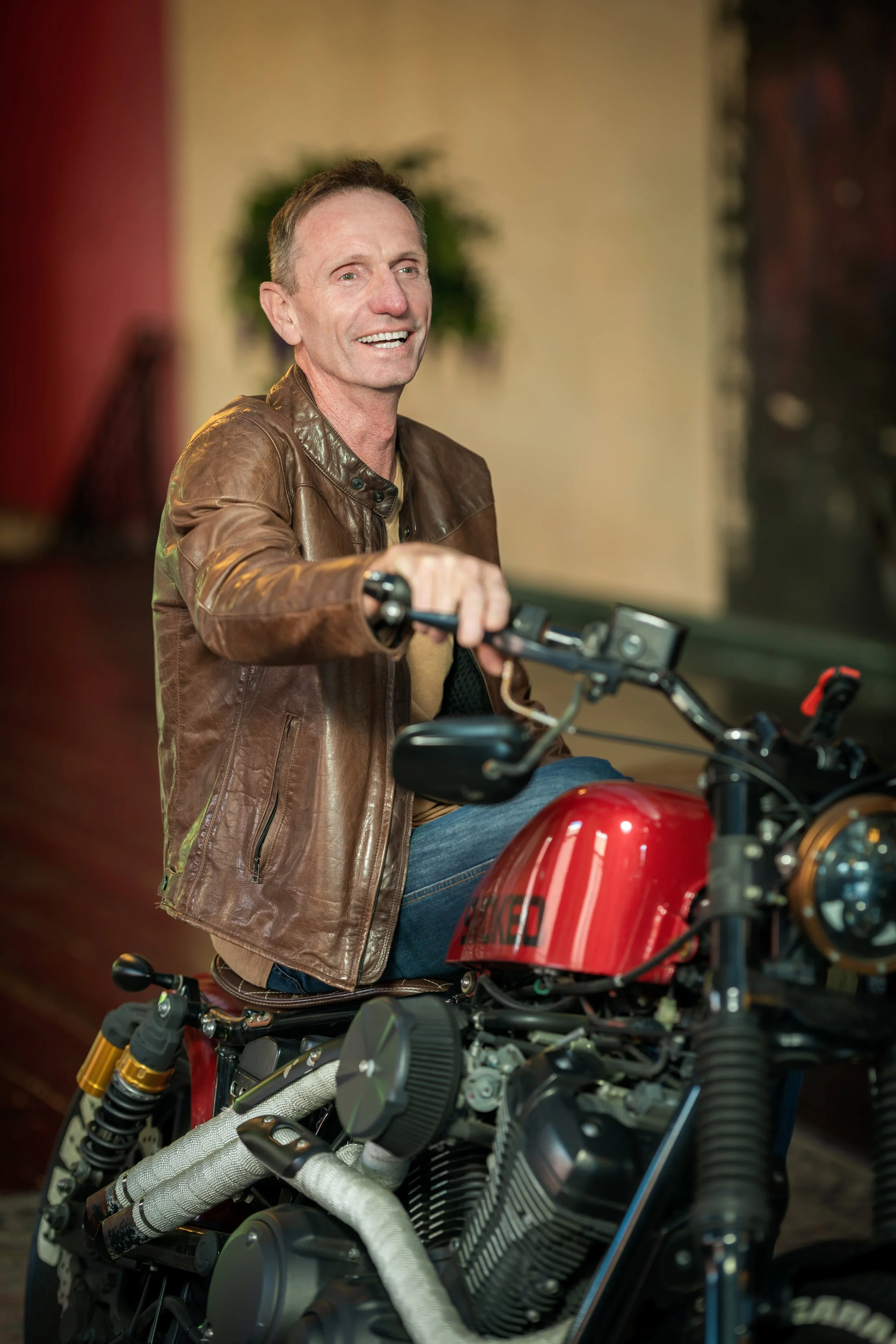 Jaye Stevens in a vintage brown leather jacket smiling while casually leaning on a red motorcycle .