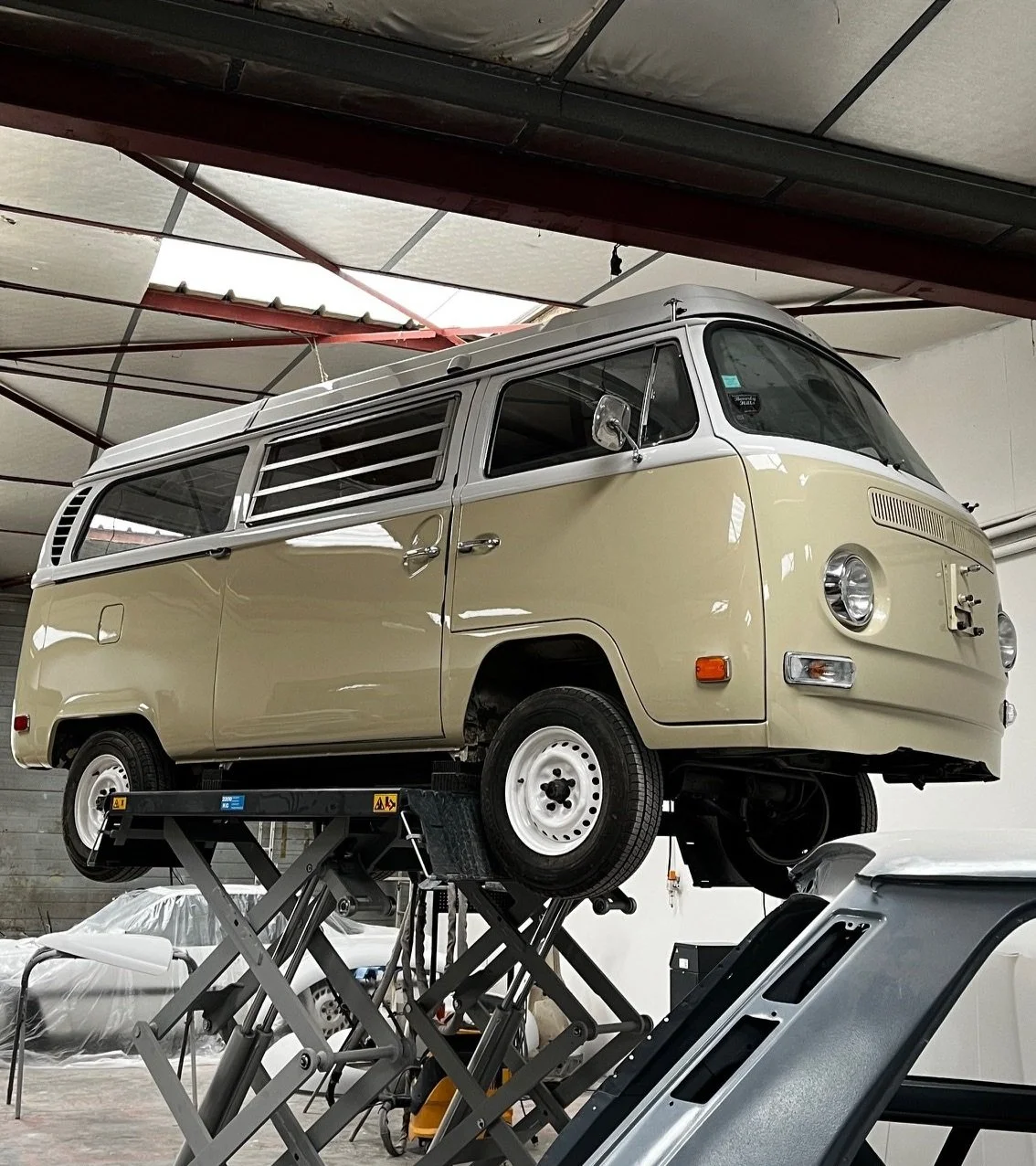 Une Volkswagen Type 2, également appelée Combi ou Microbus, en cours de restauration ou de maintenance, est levée sur un pont élévateur dans un garage ou atelier.