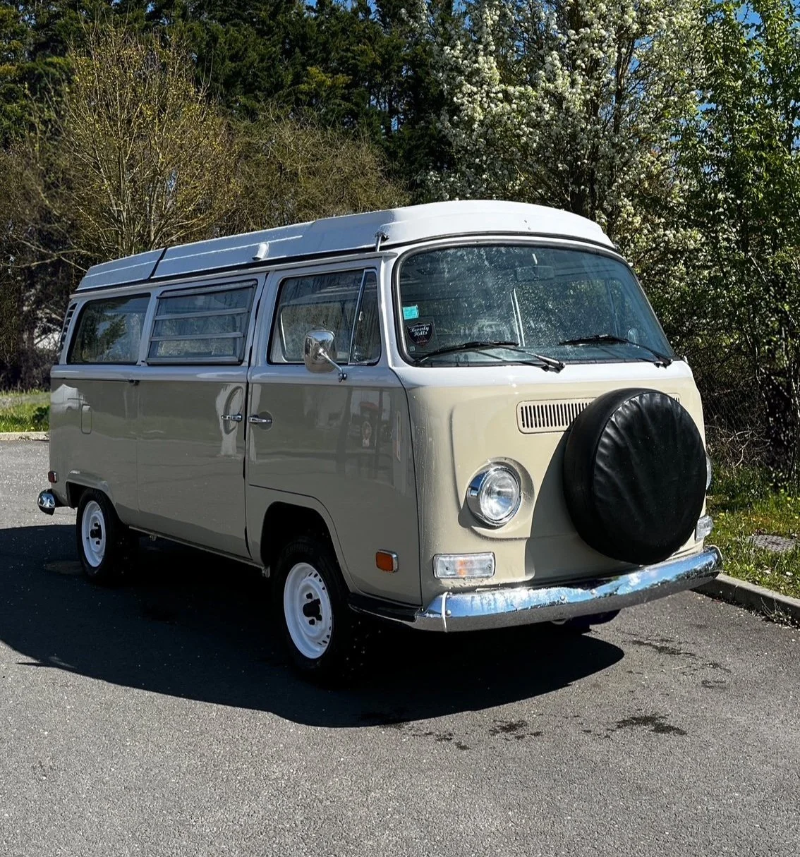 Van classique coloris beige clair avec une roue de secours recouverte d'une housse noire, stationnée sur une route en plein air, avec arrière-plan d'arbres et de ciel bleu.