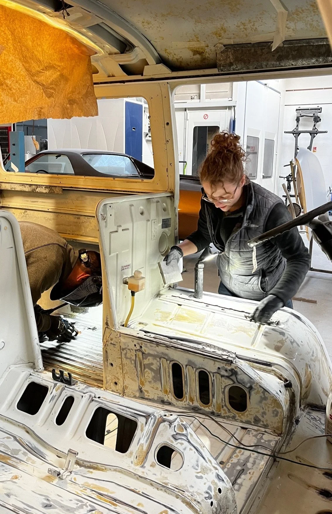 Deux personnes travaillent à la restauration d'une vieille voiture dans un atelier, l'une utilisant un outil électrique et l'autre en train de fixer quelque chose à l'intérieur du véhicule.