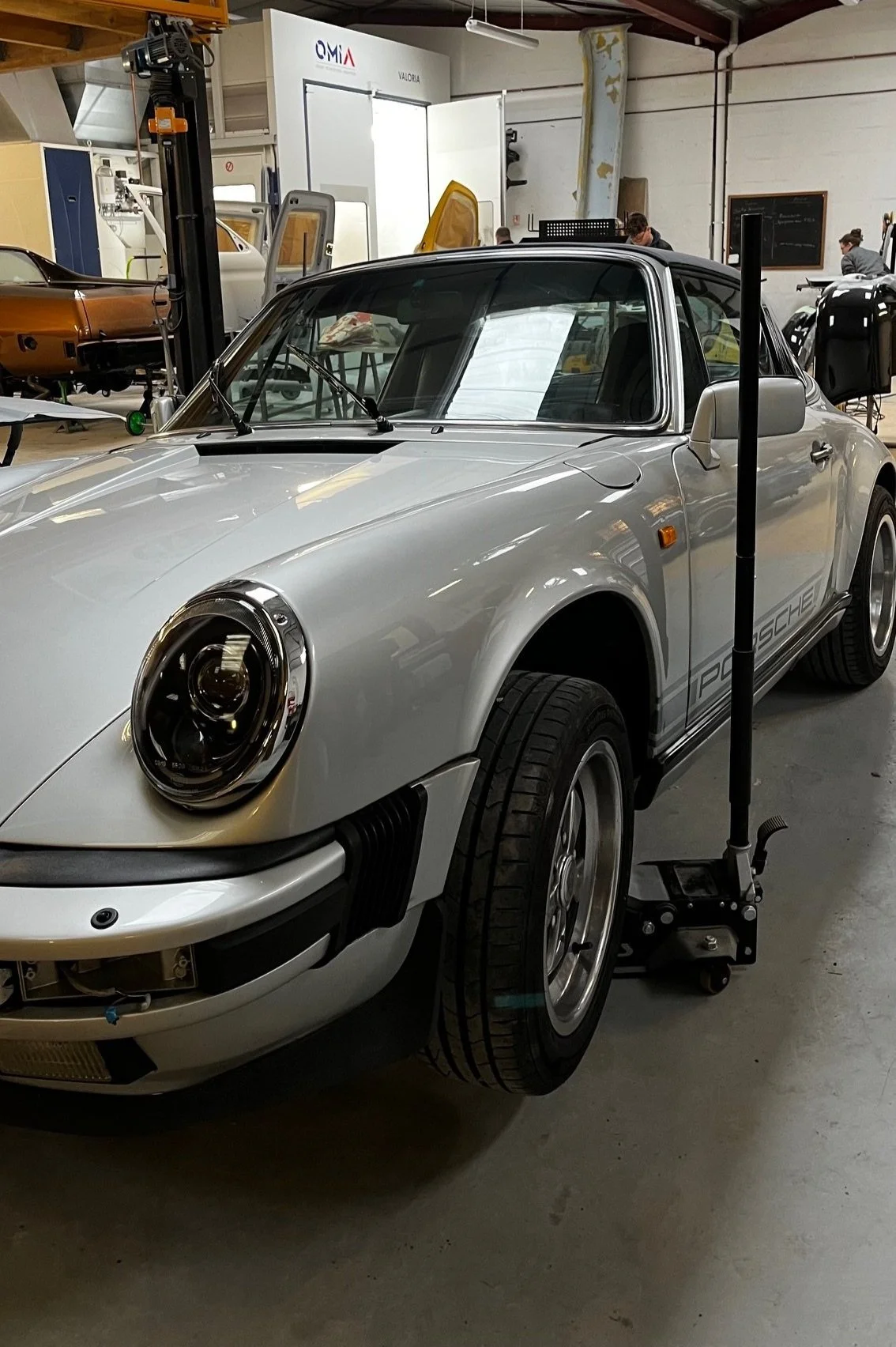 Une voiture Porsche 911 classique en cours de restauration dans un atelier. La voiture est grise avec un logo Porsche sur le côté et se trouve sur un support roulant. Il y a des outils et d'autres véhicules en arrière-plan.