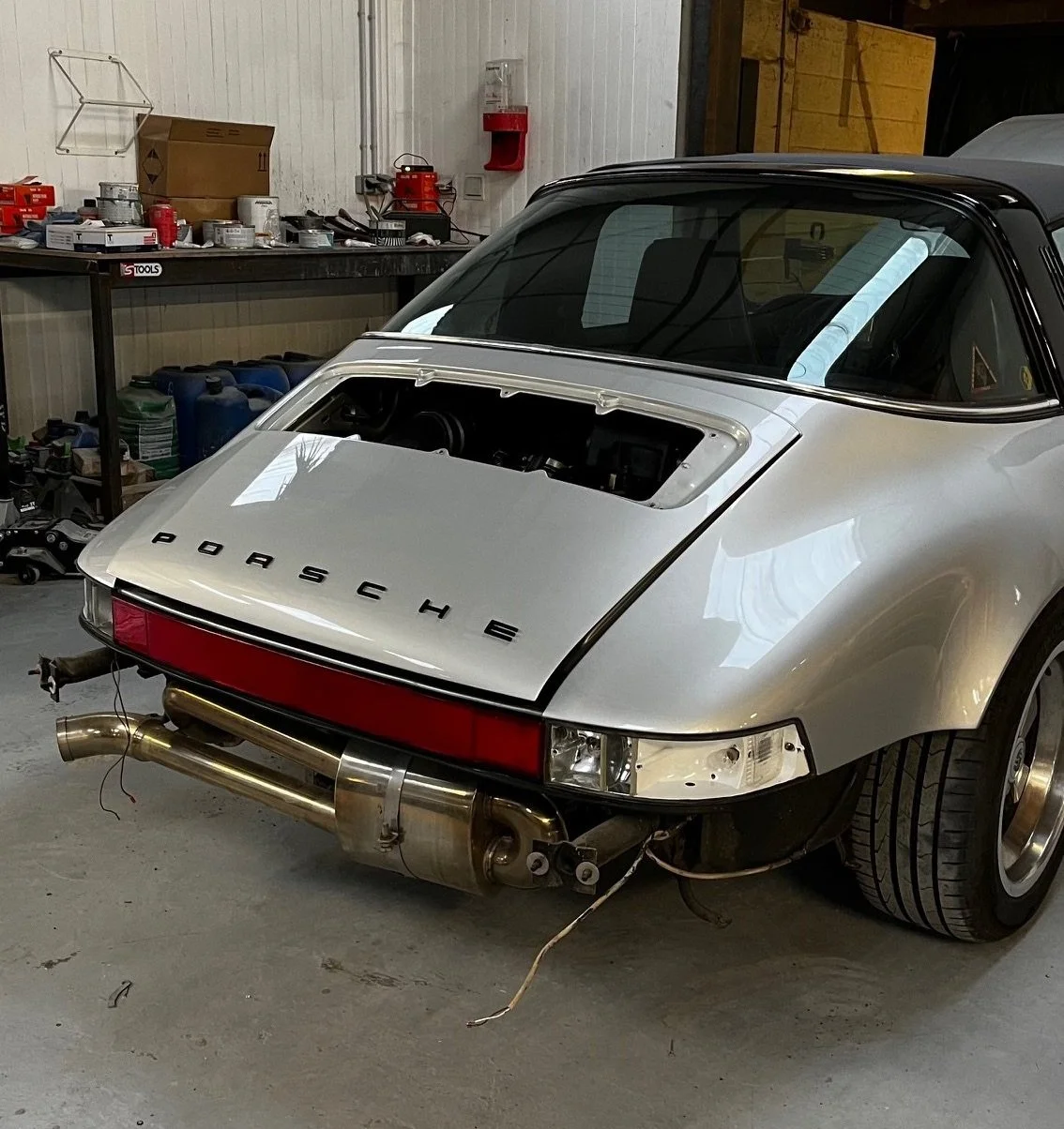 Voiture de sport Porsche vintage en cours de restauration dans un garage, sans la partie arrière du capot ouverte, avec des outils et pièces en arrière-plan.