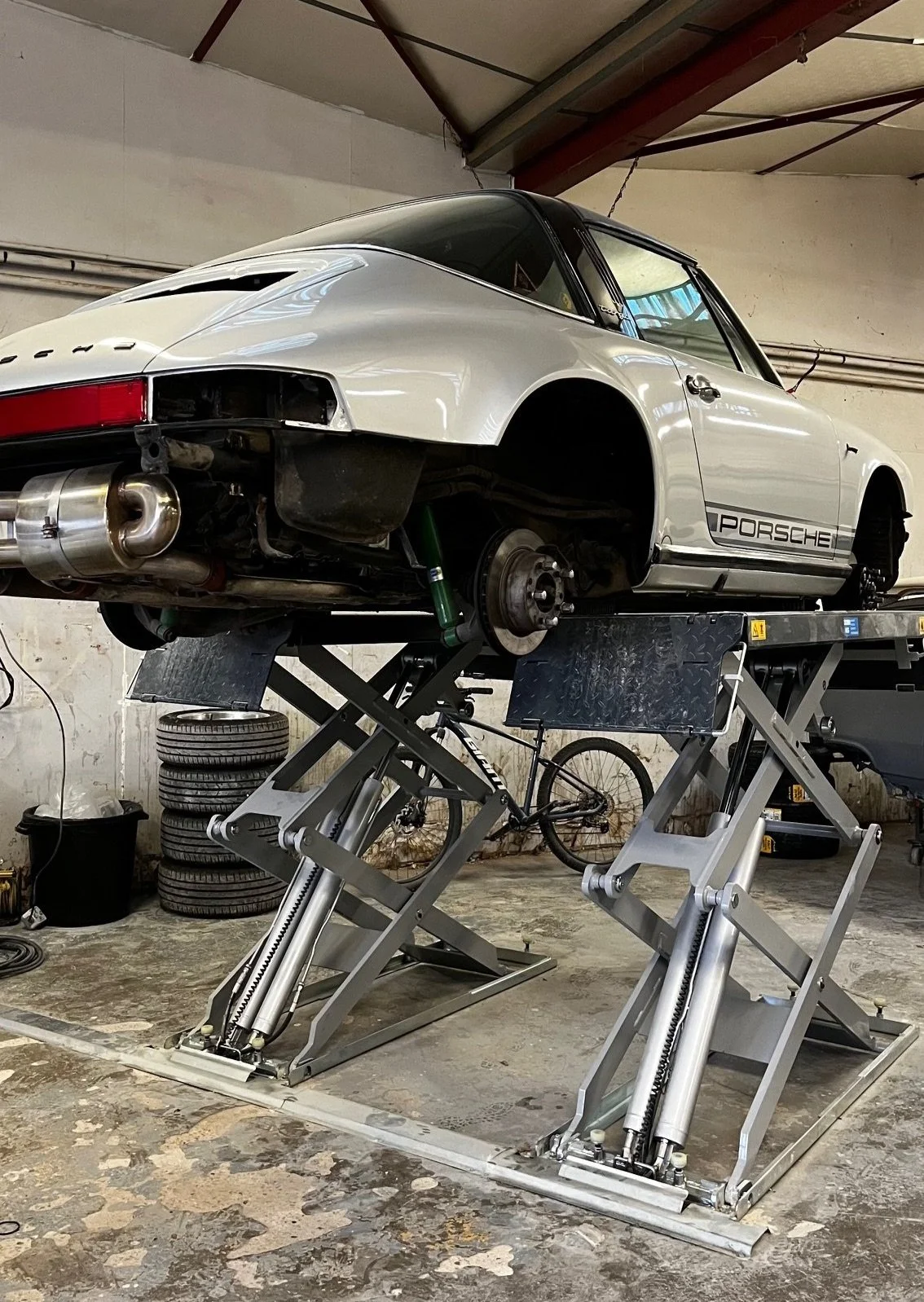Voiture Porsche grise élevée sur un support de maintenance dans un garage, sans roues, avec outils et pneus empilés en arrière-plan.