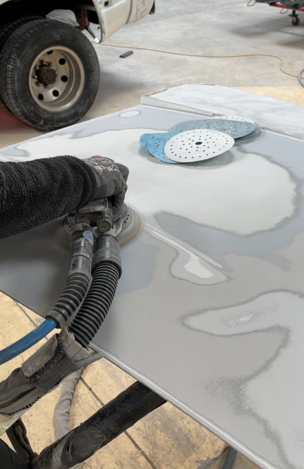 Une personne utilise une ponceuse sur un capot pour la préparation, avec des disques abrasifs posés sur la surface à côté. Un véhicule est visible en arrière-plan dans un atelier ou garage.
