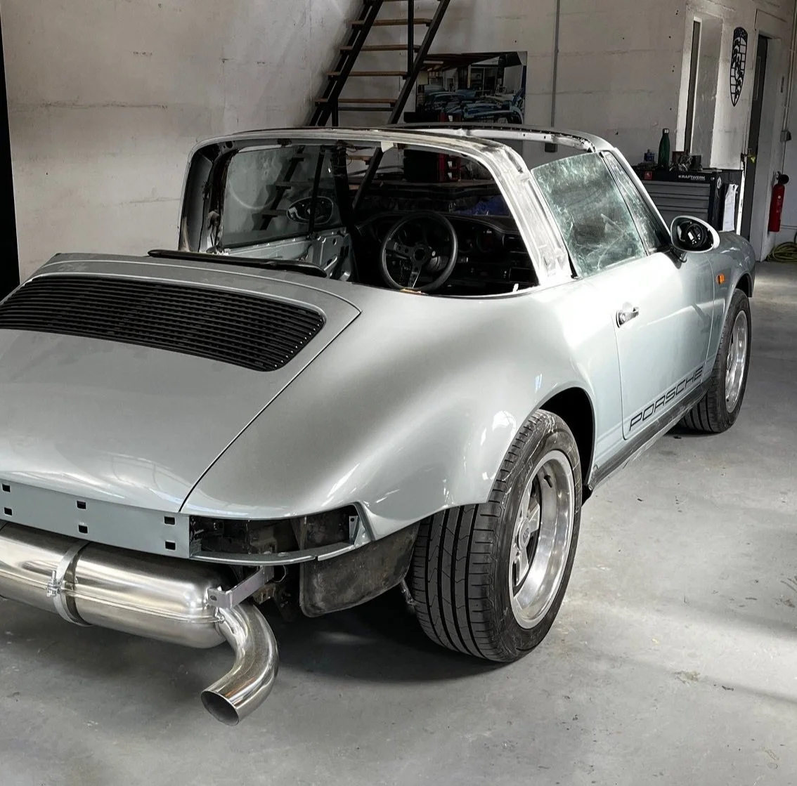 Voiture Porsche classique en cours de restauration, stationnée dans un garage, sans pare-chocs avant, avec un moteur équipé d'un échappement large en aluminium.