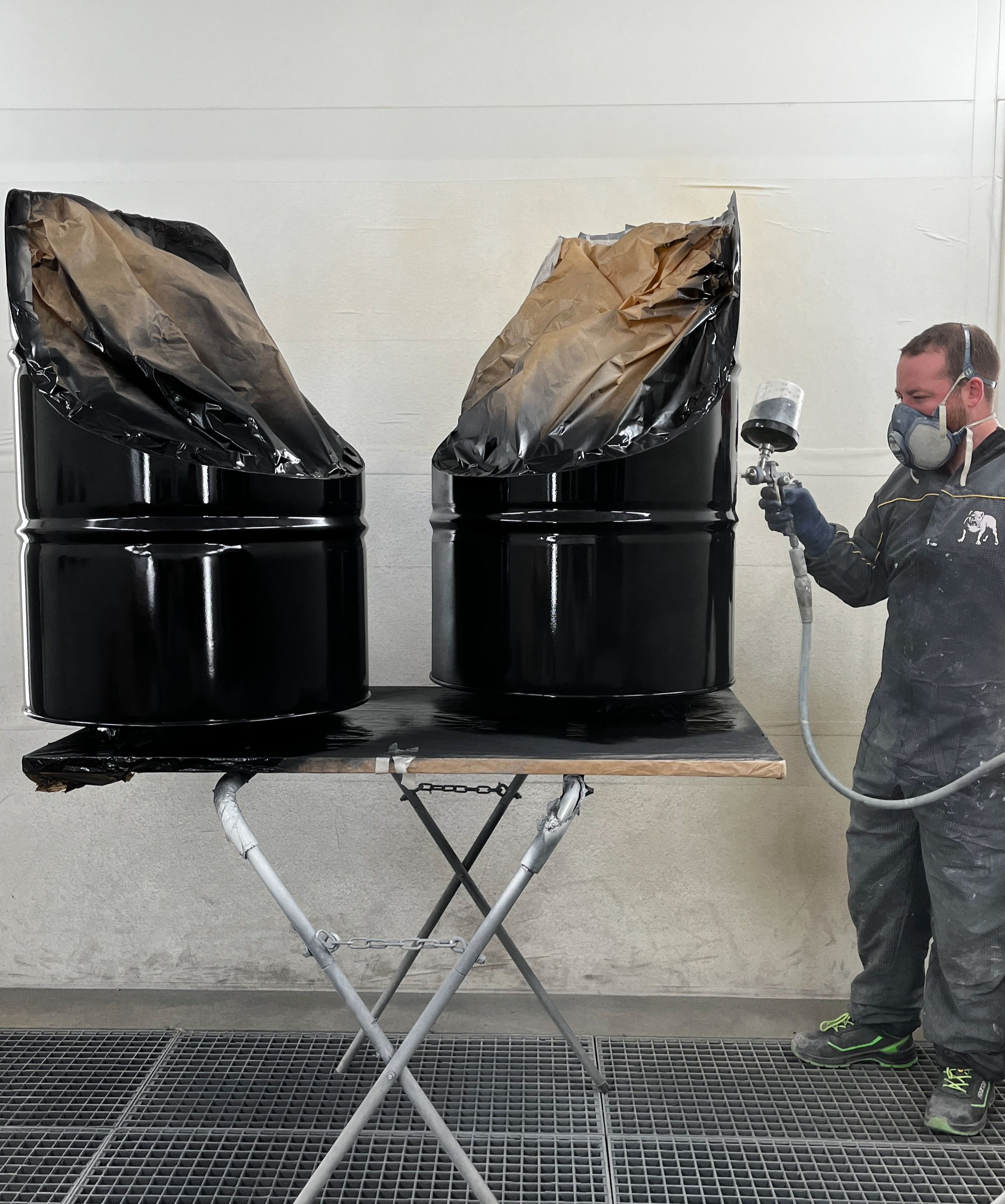 Un homme en combinaison de protection utilise un pistolet à peinture pour peindre deux barils métalliques noirs dans une cabine de peinture.