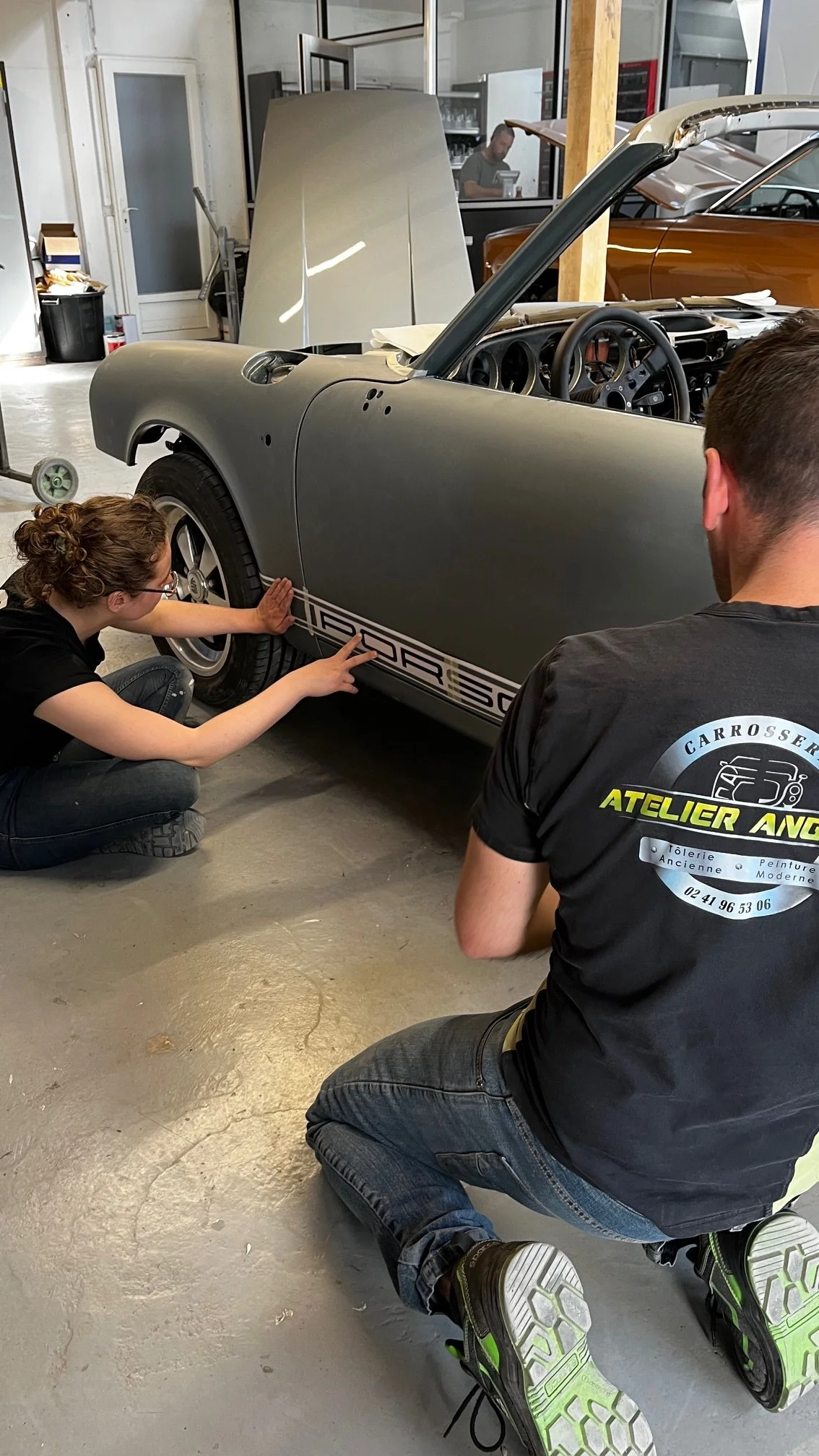 Deux personnes travaillent sur une voiture de sport en cours de restauration ou de personnalisation dans un atelier, discutant devant la porte d'une voiture de couleur grise.