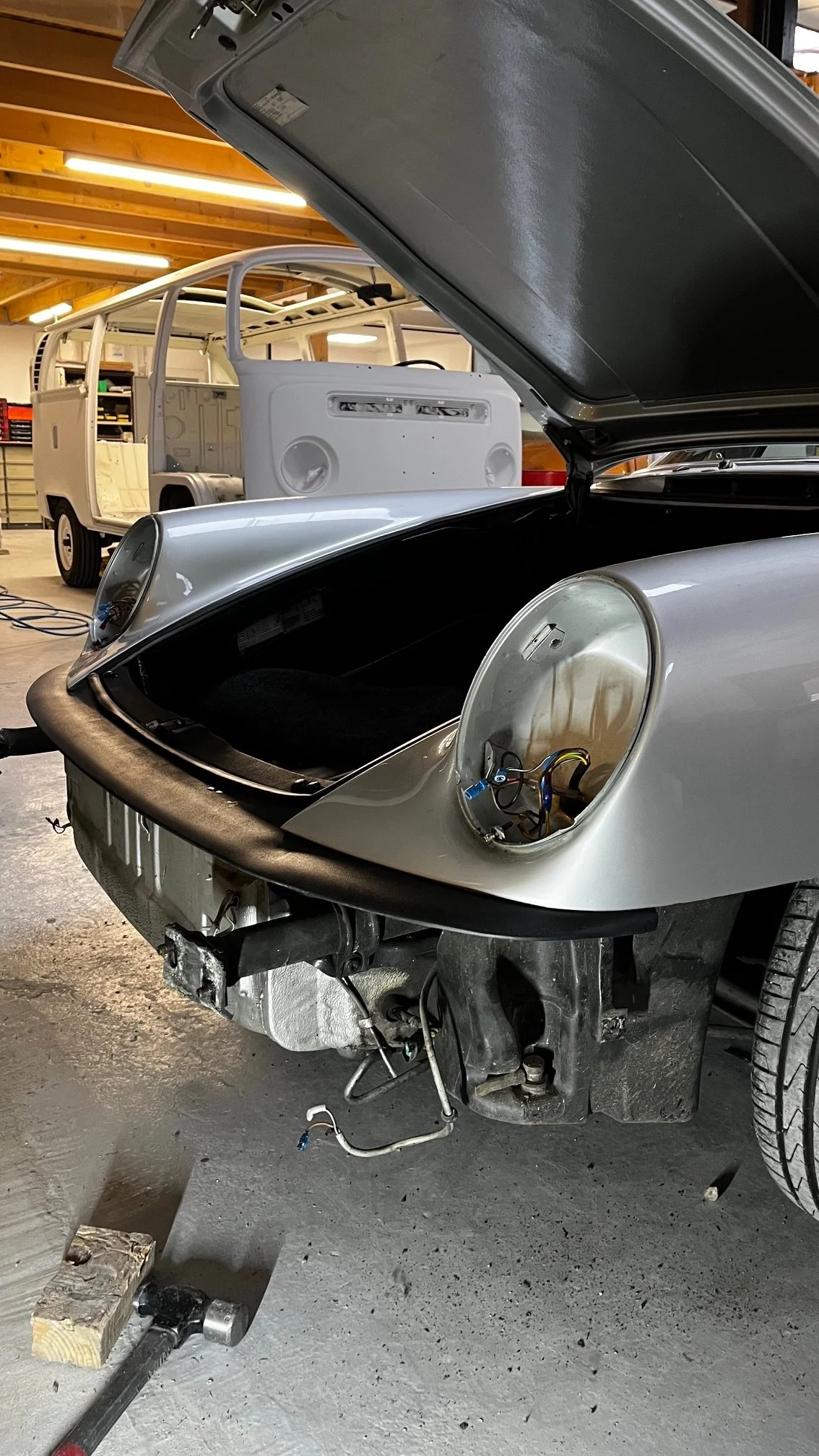 Une voiture de sport en cours de restauration ou de réparation dans un atelier, avec le capot ouvert et des composants électriques visibles, ainsi qu'une autre voiture en arrière-plan.