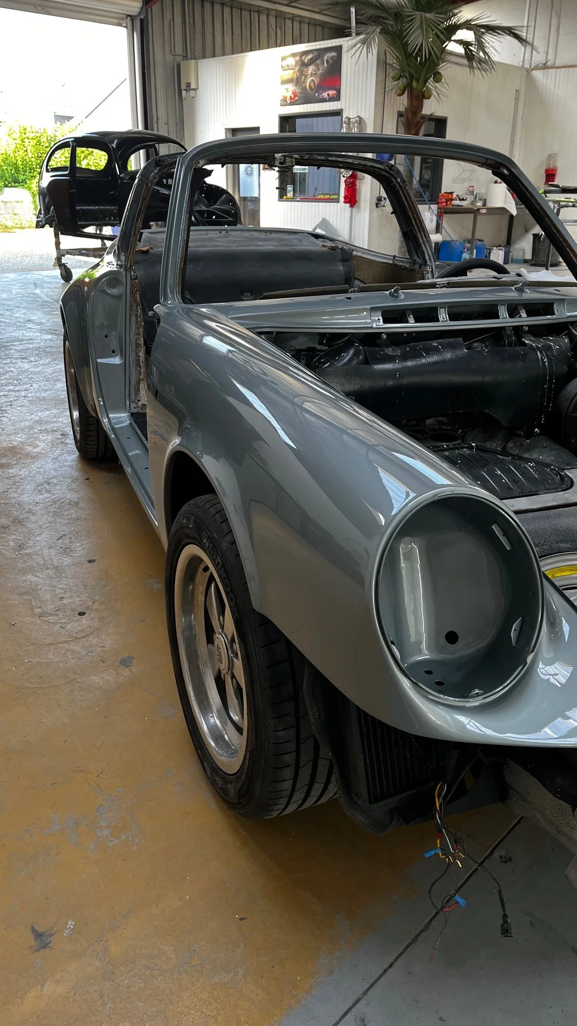 Un voiture de course en cours de restauration dans un atelier, sans phares ni moteur installé, avec une carrosserie grise et des pneus slicks.