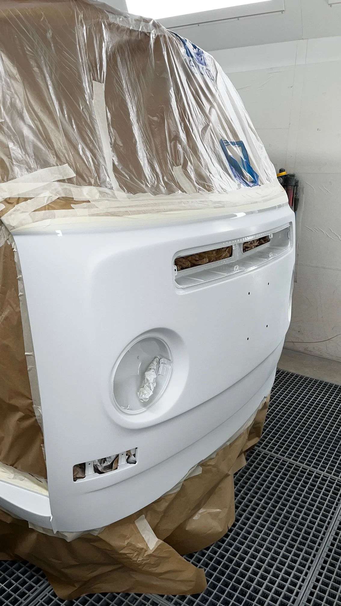 Un combi Volkswagen en cours de peinture, blanche, dans une cabine de peinture automobile, avec du papier de masquage et des protections en plastique autour.
