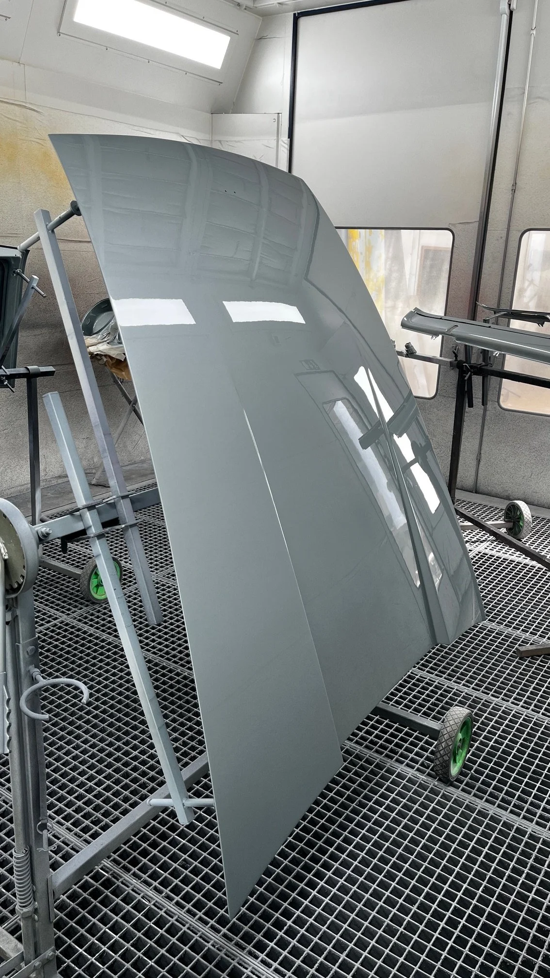 capot de voiture peint dans un atelier, posé sur un support mobile. La surface est lisse et glossy, reflétant la lumière et la structure de l'atelier.