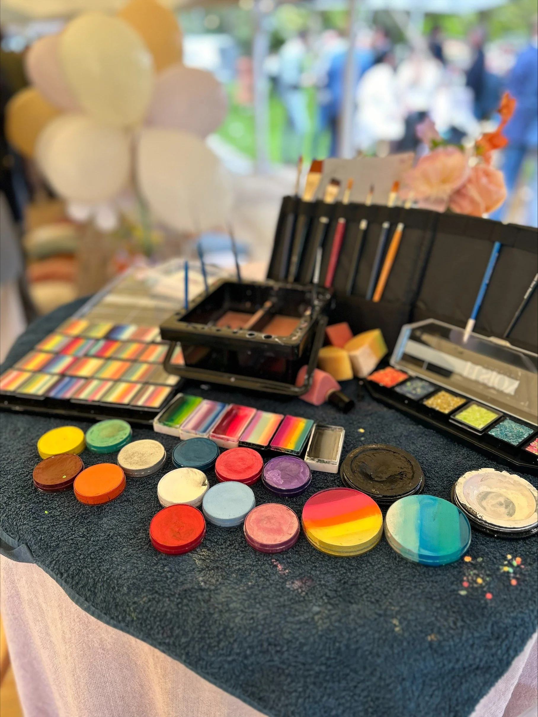 Display of face paints and makeup tools on a table at an outdoor event, with blurred people and balloons in the background.