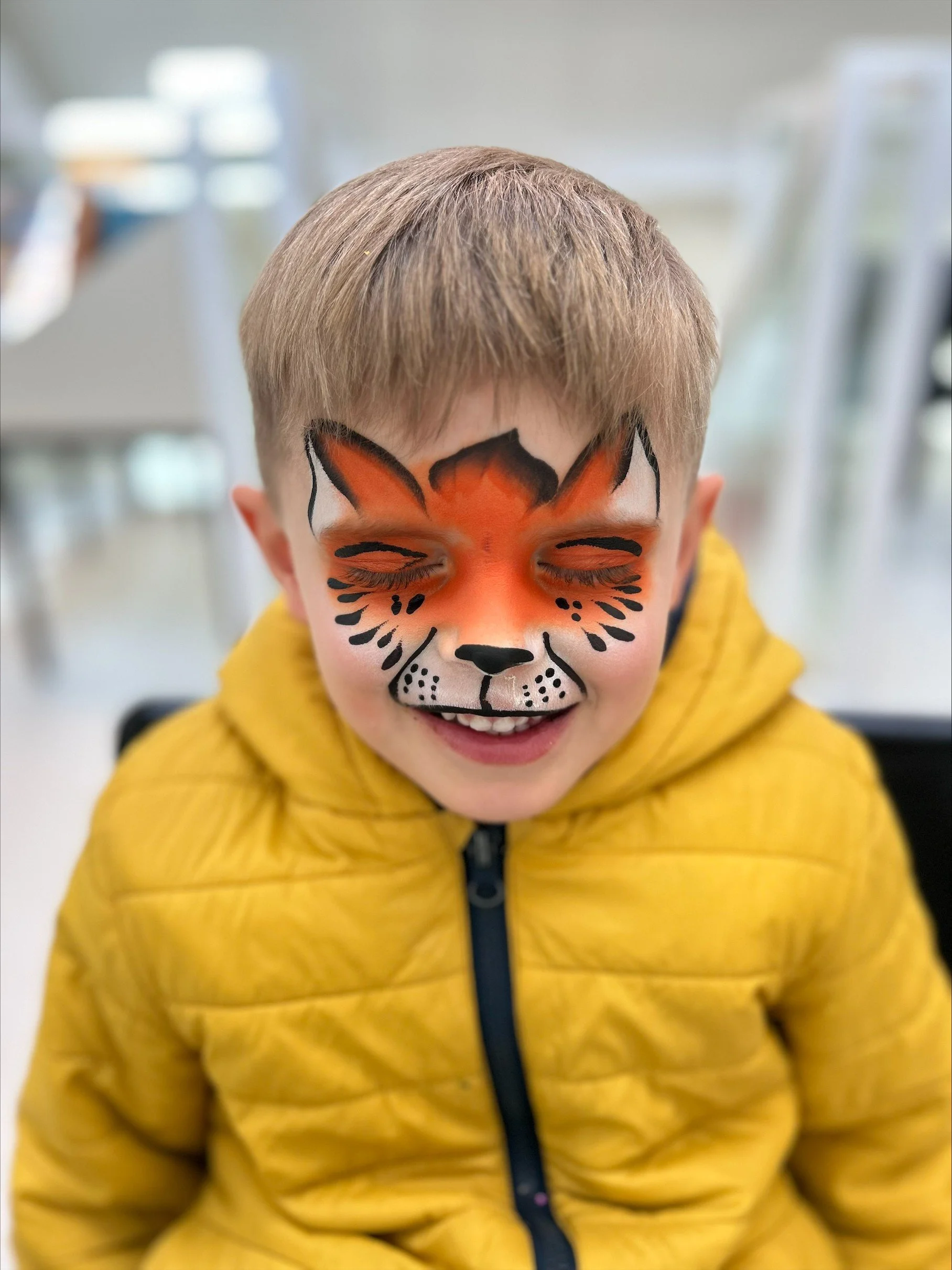 A young boy with face paint of a fox, wearing a yellow jacket, smiling with eyes closed.