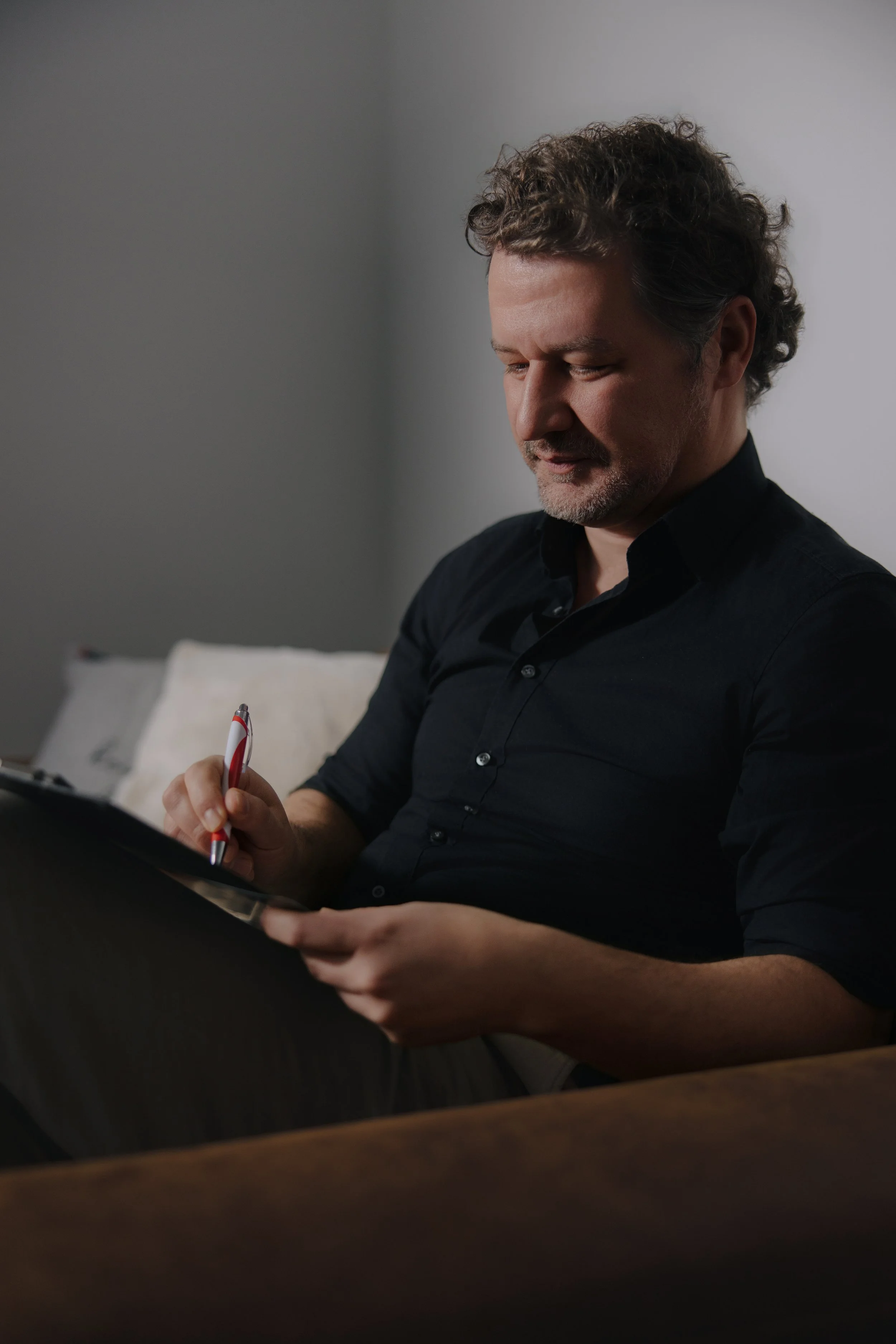 Ein Mann sitzt auf einem Sofa, liest vielleicht einen Zettel oder ein Tablet, hält einen roten Kugelschreiber.