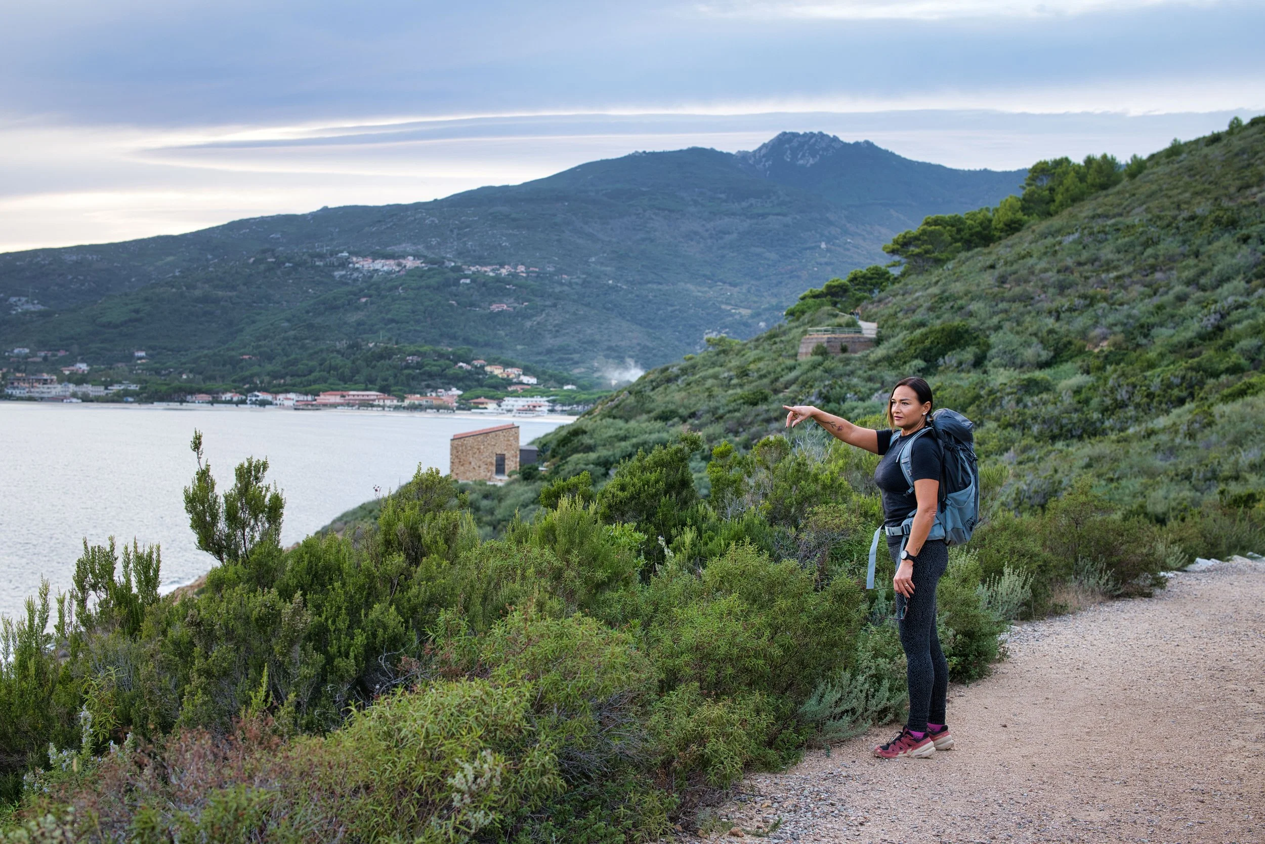 Una donna con zaino punta il paesaggio che comprende montagne, acqua e vegetazione durante una passeggiata nella natura.