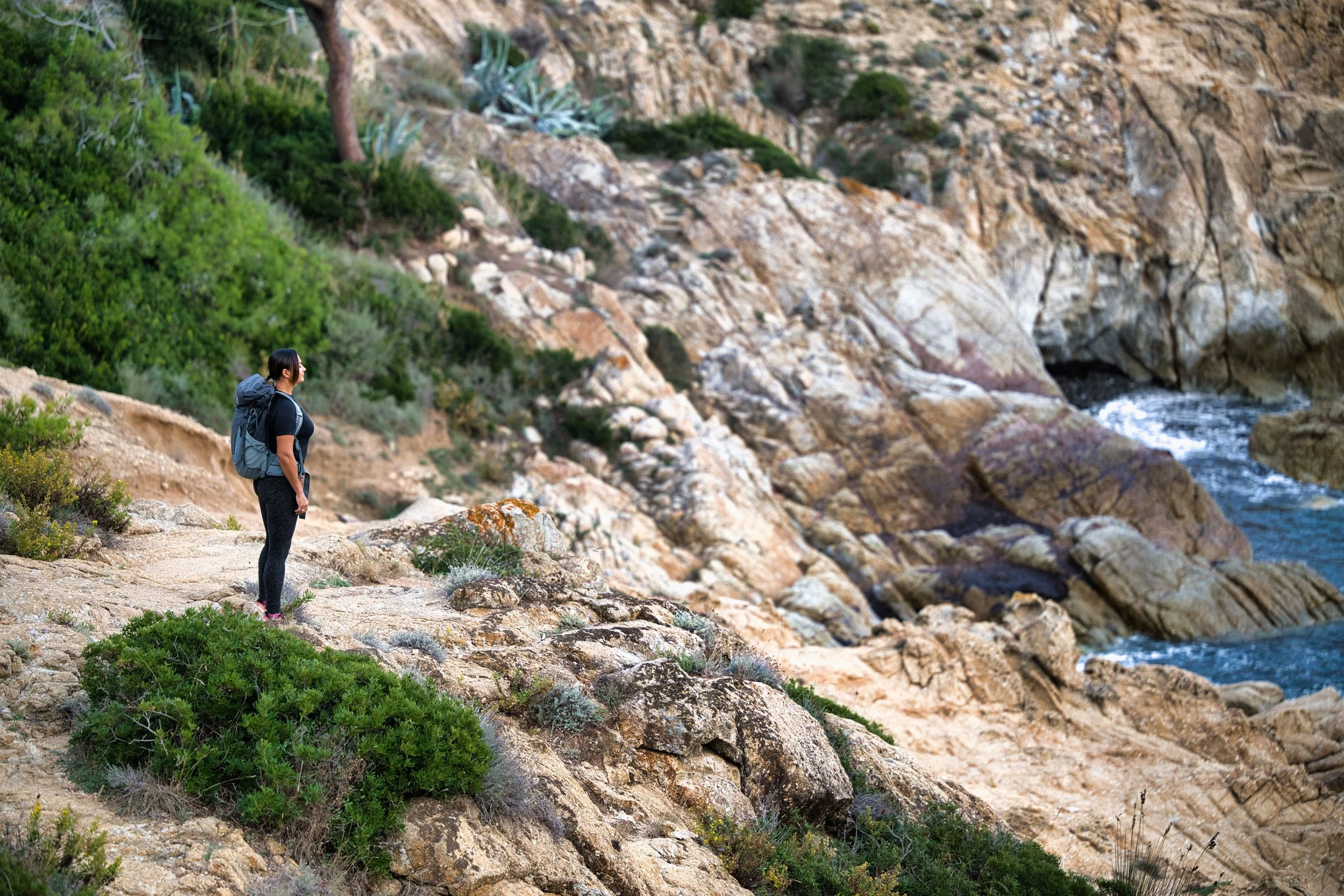 Una donna con zaino che fa trekking su una scogliera vicino al mare con rocce e vegetazione.