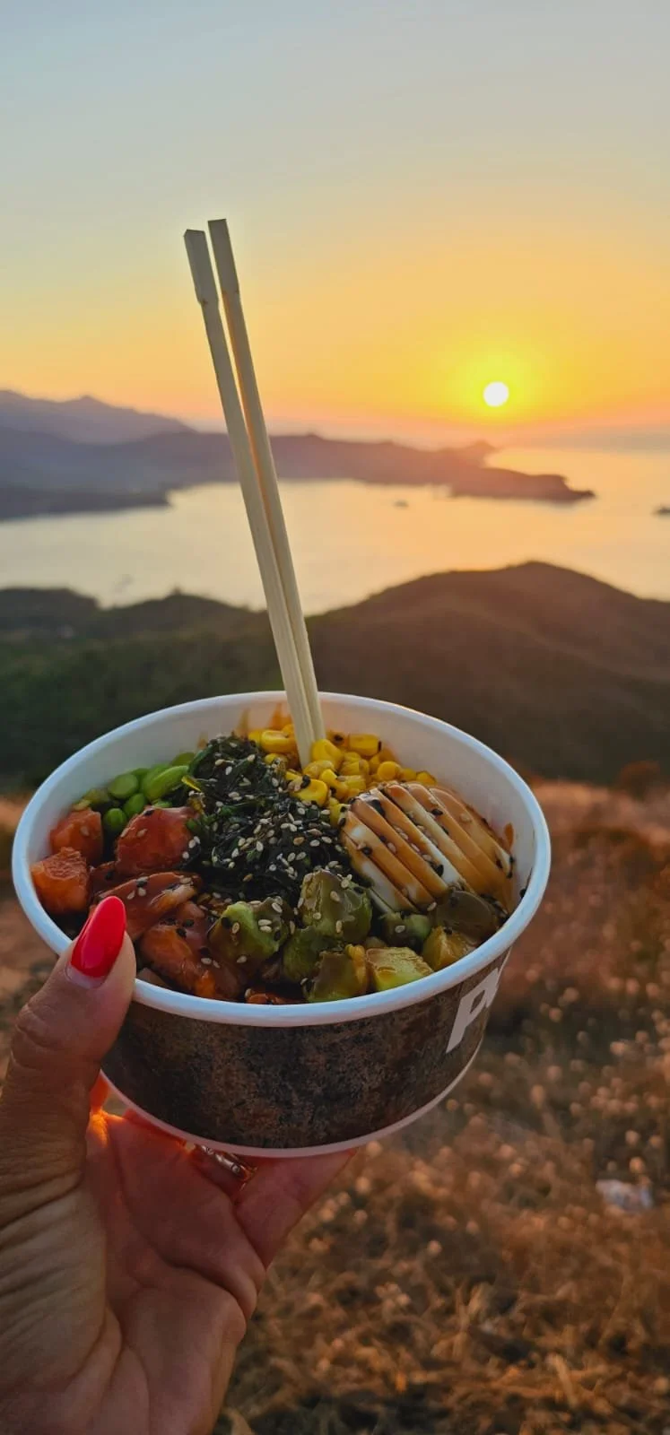 Ci crede una ciotola di poke con verdure, semi di sesamo e pesce, con uno sfondo di tramonto su un paesaggio di montagna e lago.