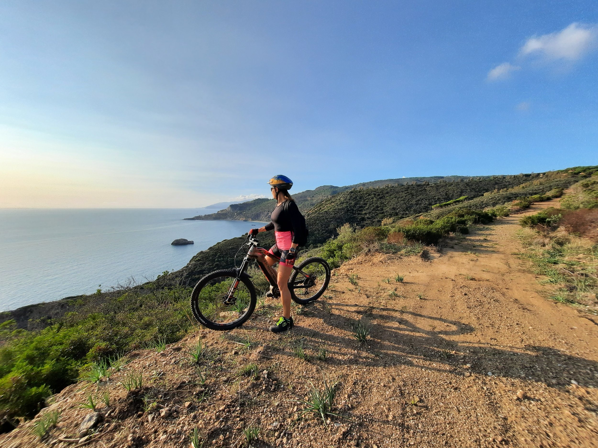 Una donna con casco e abbigliamento da ciclismo si ferma con la sua bicicletta sulla strada sterrata vicino alla costa con vista sull'oceano e le colline verdeggianti.