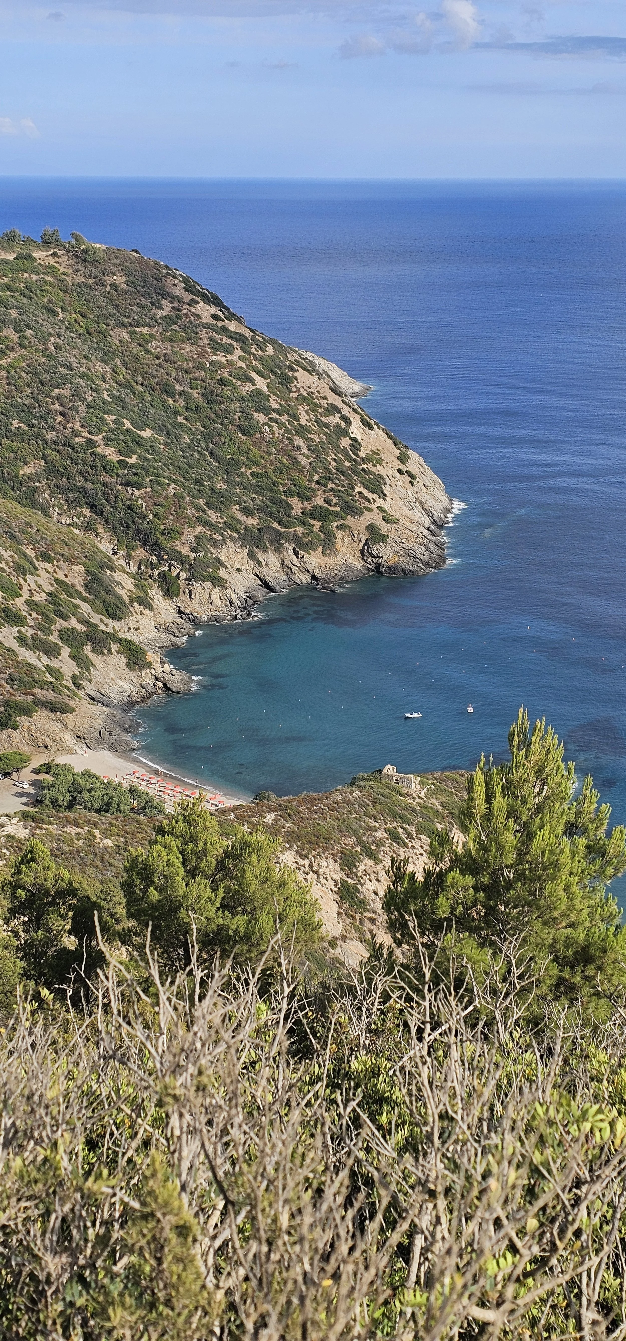 Paesaggio costiero con spiaggia, scogli e cielo azzurro, vista dall'alto di una baia circondata da colline verdi e arbusti.