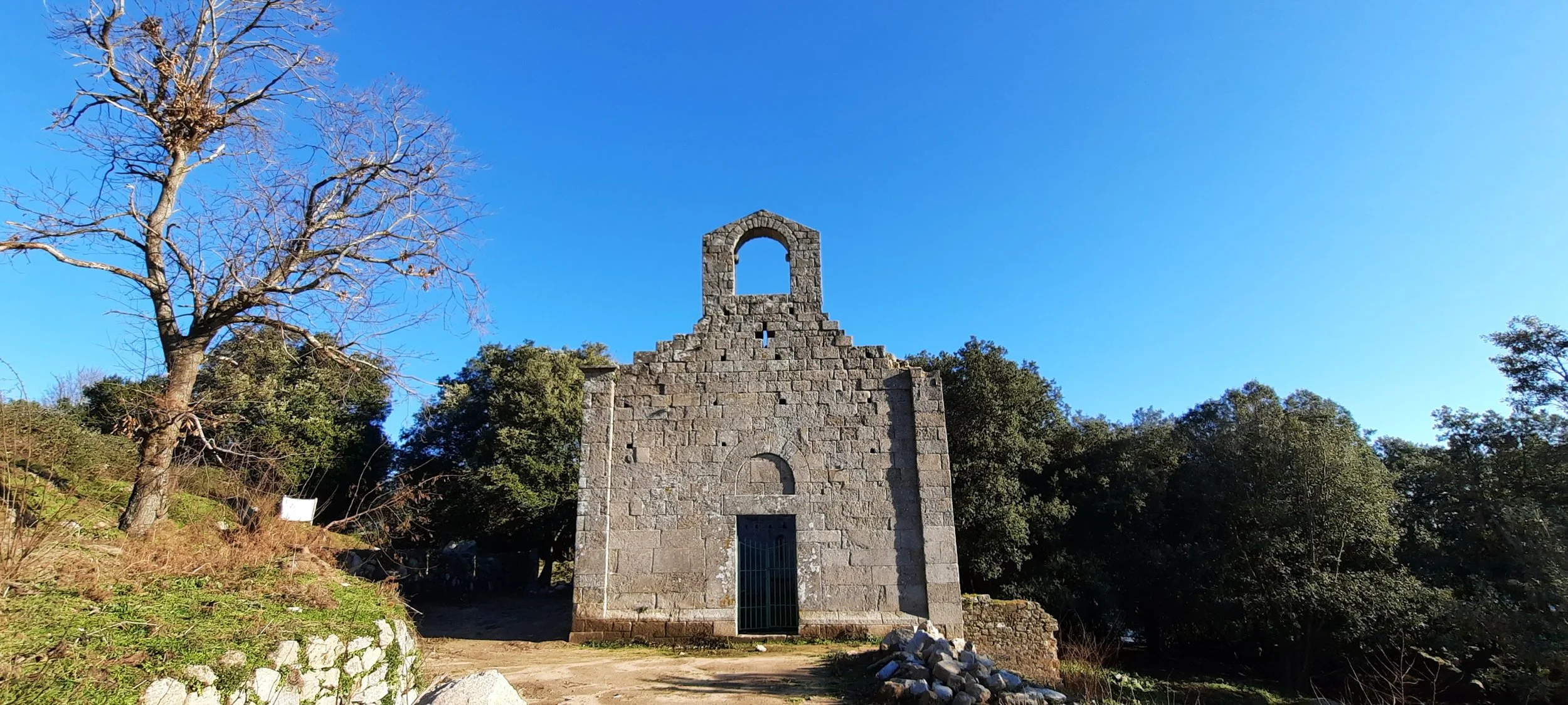 Resti di una chiesa di pietra con archi e pareti rovinate, circondata da alberi e natura, sotto un cielo blu.
