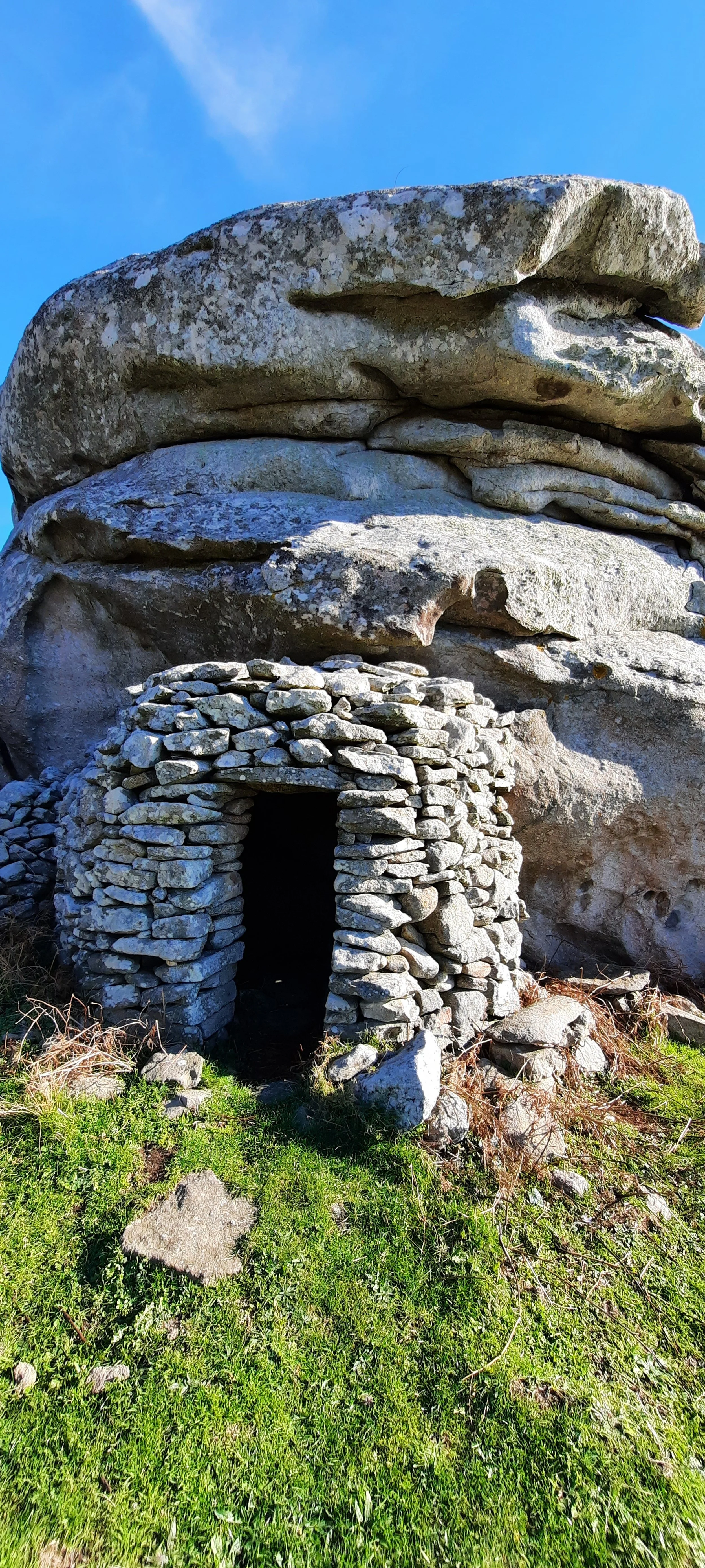 Struttura di pietre disposte a formare un'entrata di un riparo o un ricovero sotto grandi rocce, con erba e pietre sul terreno circostante, sotto un cielo azzurro.