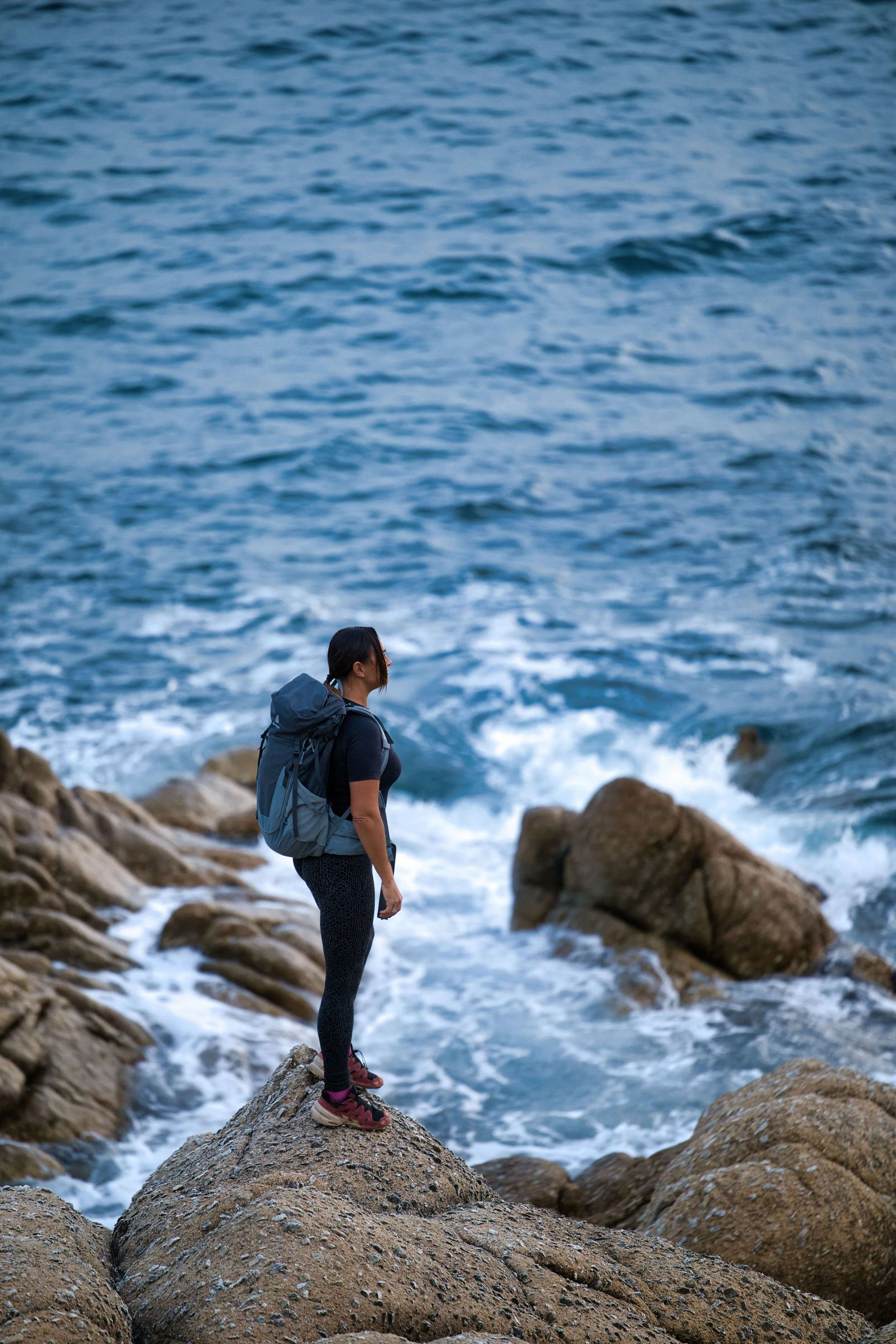 Donna con zaino che si trova su una roccia vicino al mare.