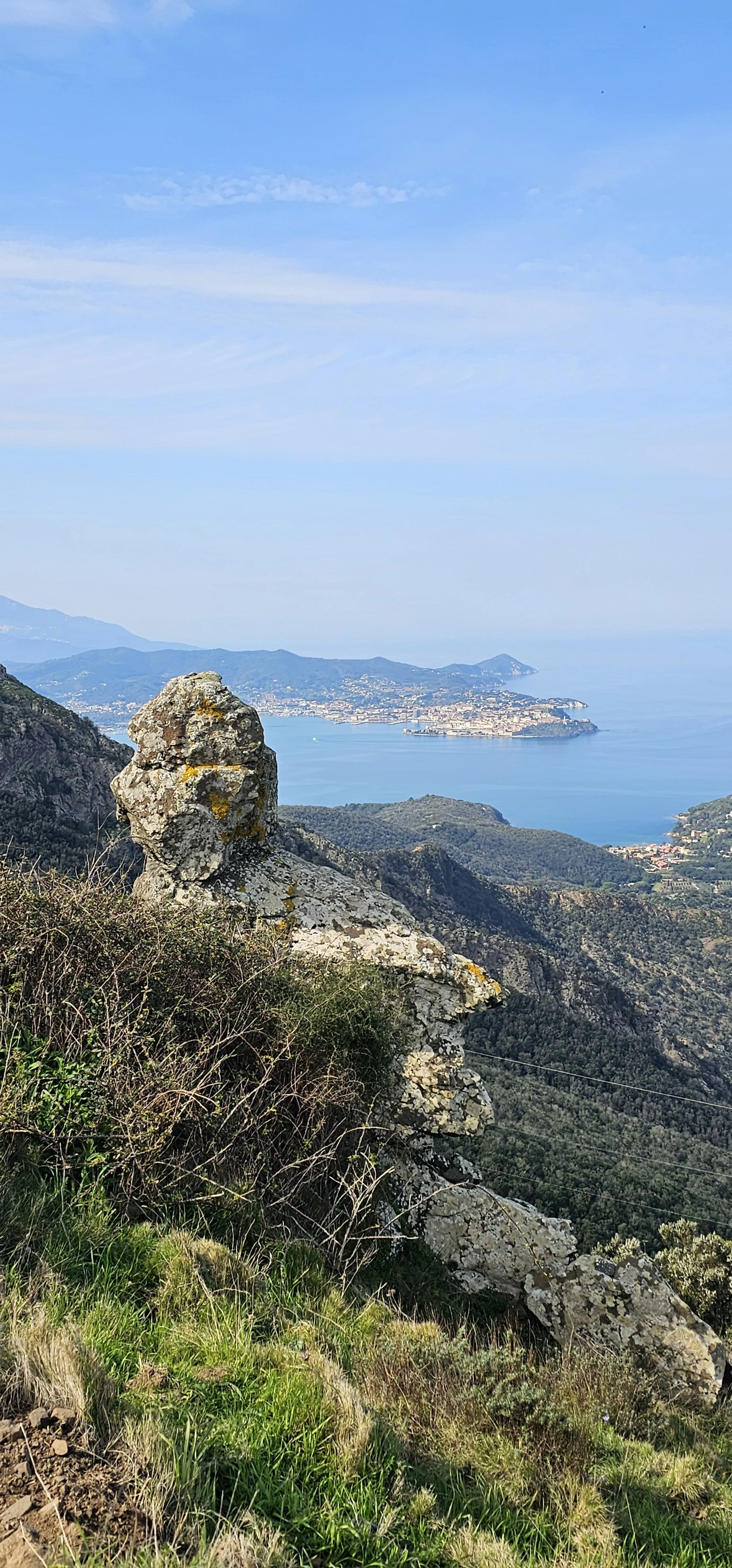 Paesaggio naturale con rocce e vegetazione in primo piano, vista sul mare e città sullo sfondo, cielo sereno.
